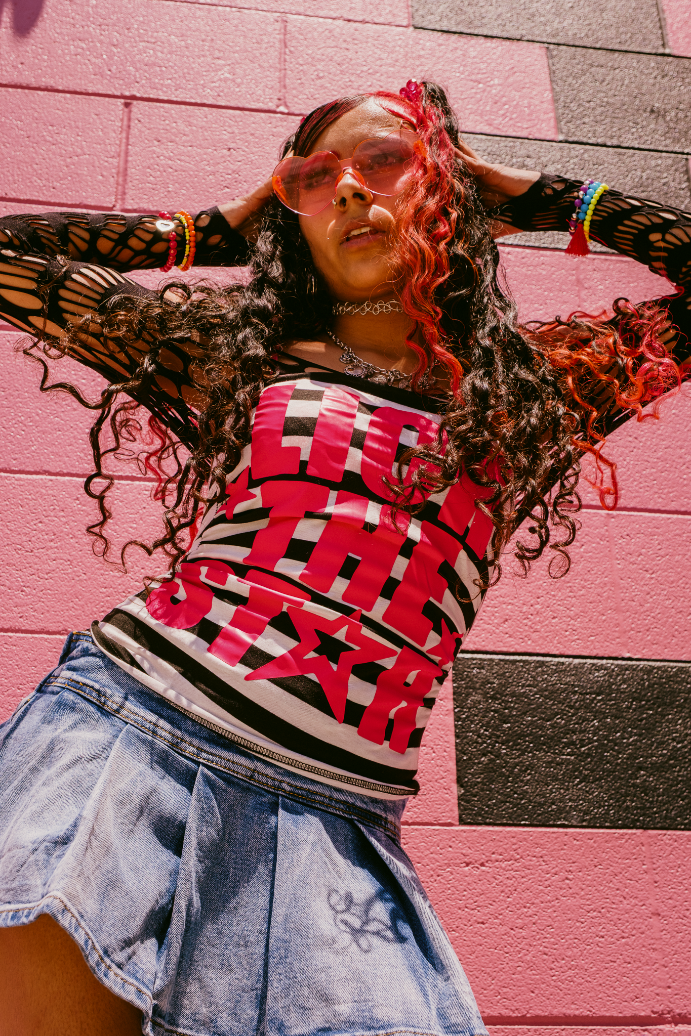 Young woman in red graphic top, denim skirt, fishnet sleeves, and pink heart sunglasses posing against a pink brick wall.