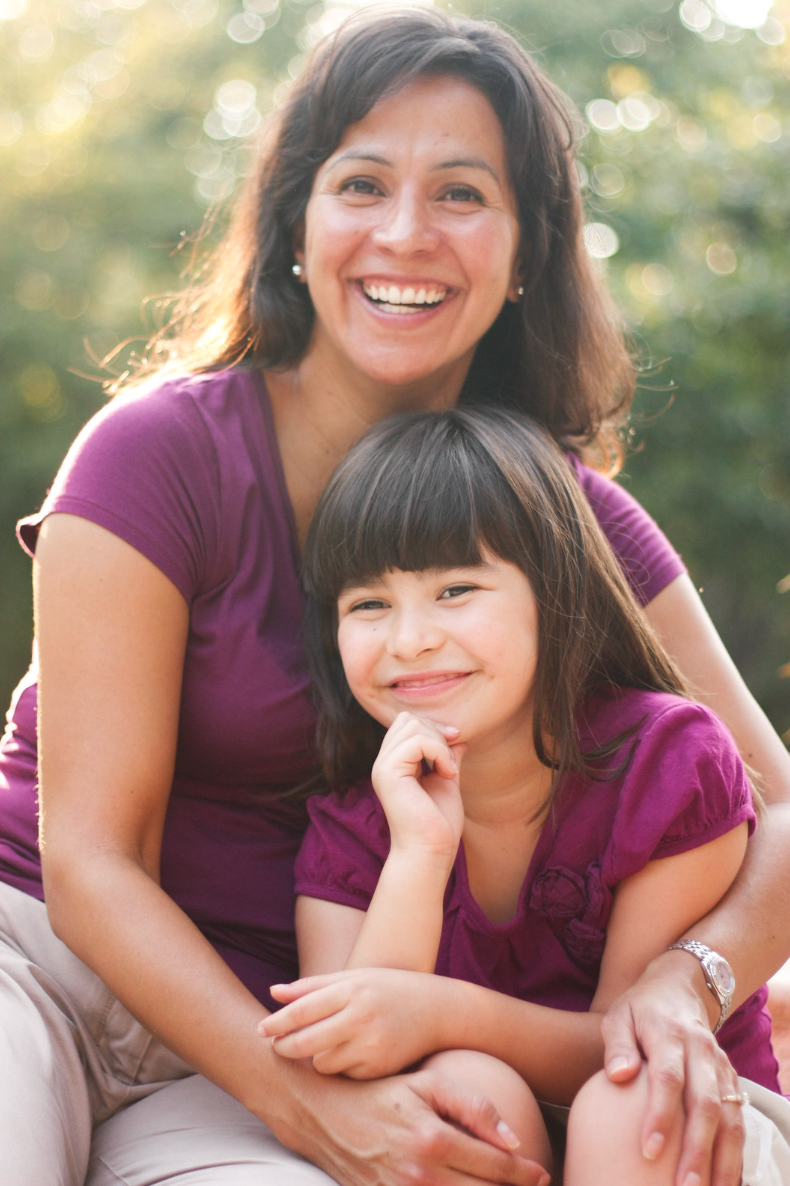 Smiling Hispanic mother and daughter in matching purple shirts posing outdoors in warm sunlight.