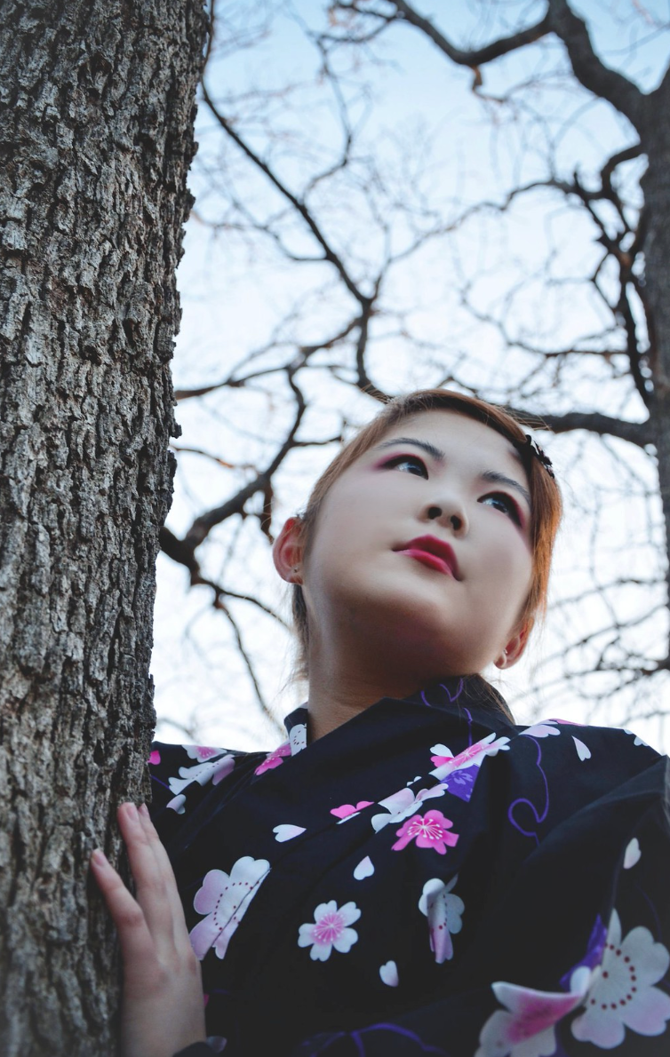 Young Asian woman in floral kimono leaning against a tree trunk looking upward.