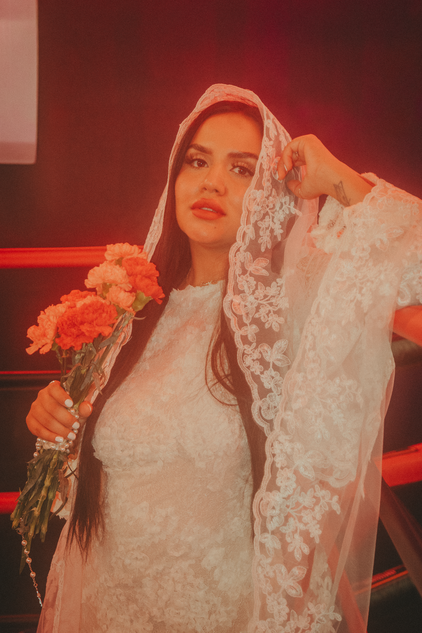 Bride in white lace dress and veil holds pink carnations under red ambient lighting.