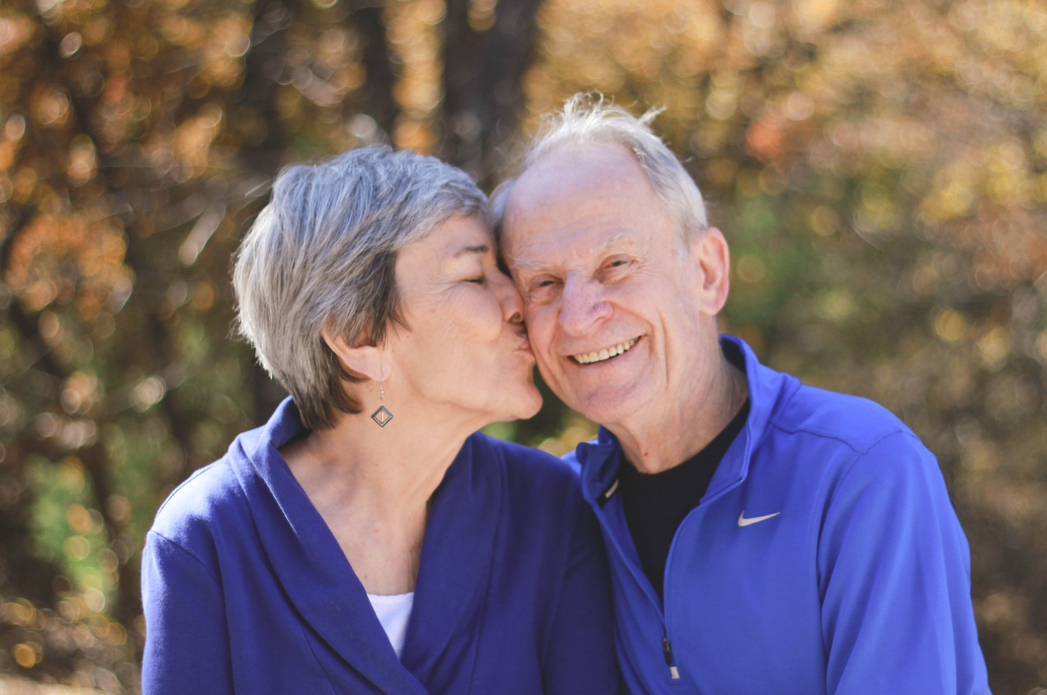 Senior couple in blue jackets sharing a kiss outdoors among autumn foliage.