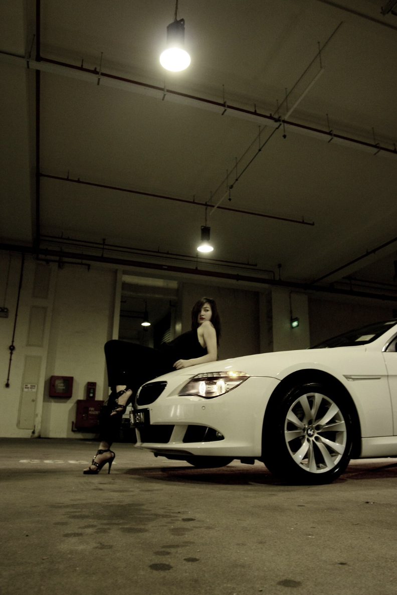 Woman posing beside a white BMW in a dimly lit parking garage.