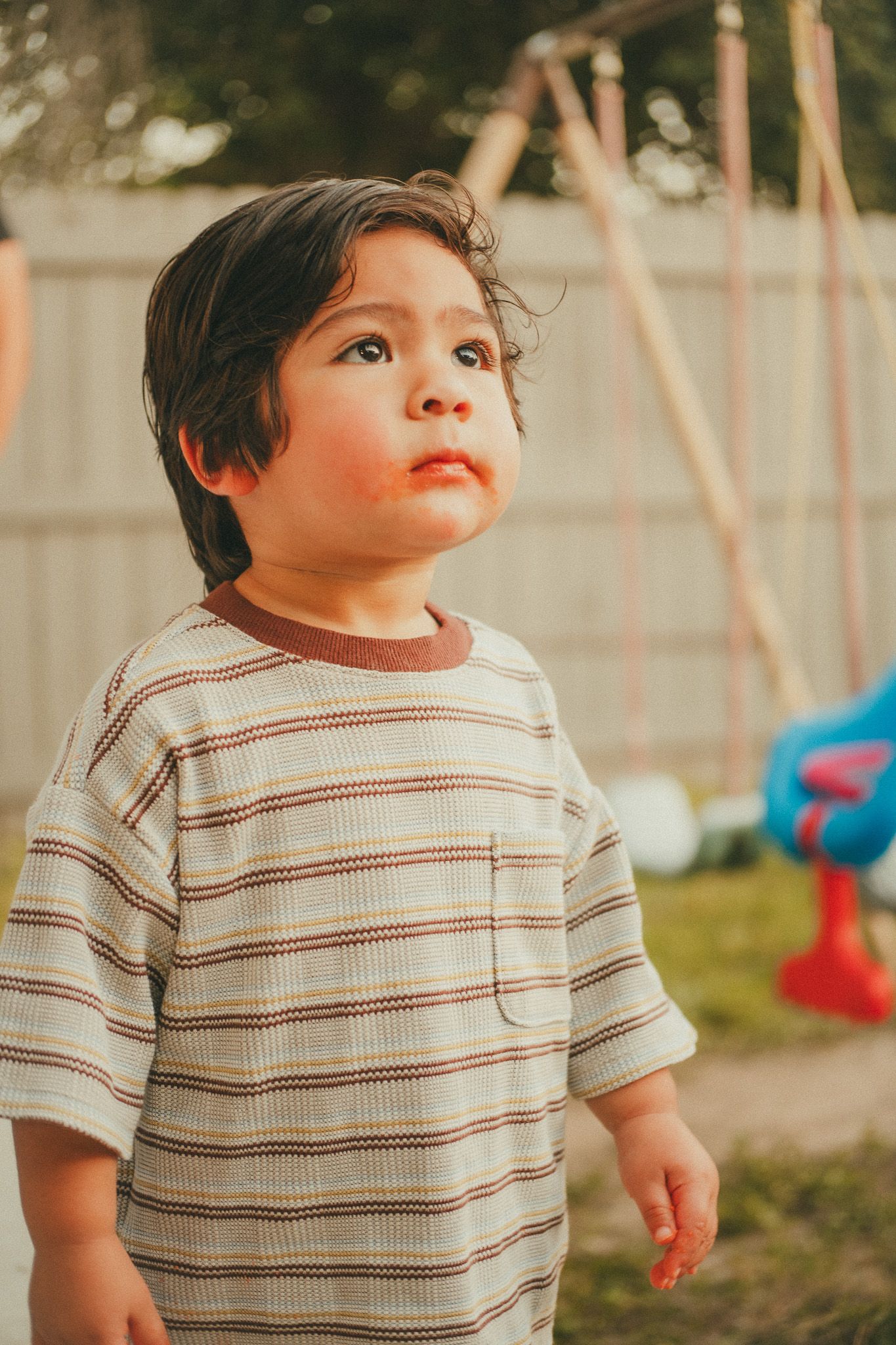 Young toddler boy in striped shirt standing in backyard near swing set, looking up curiously.