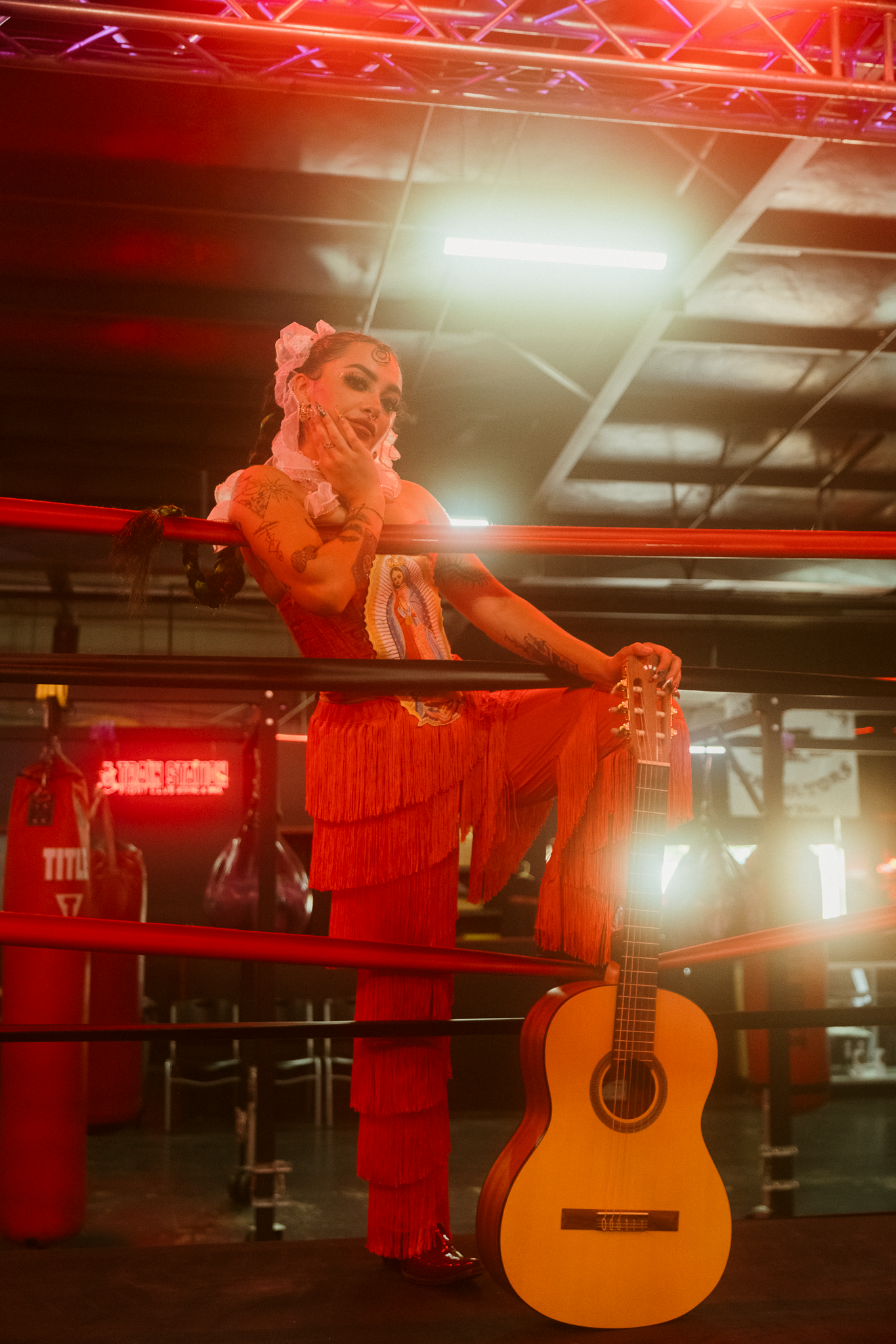 A performer in red fringe costume with skull makeup poses in a boxing ring beside an acoustic guitar under red lighting.