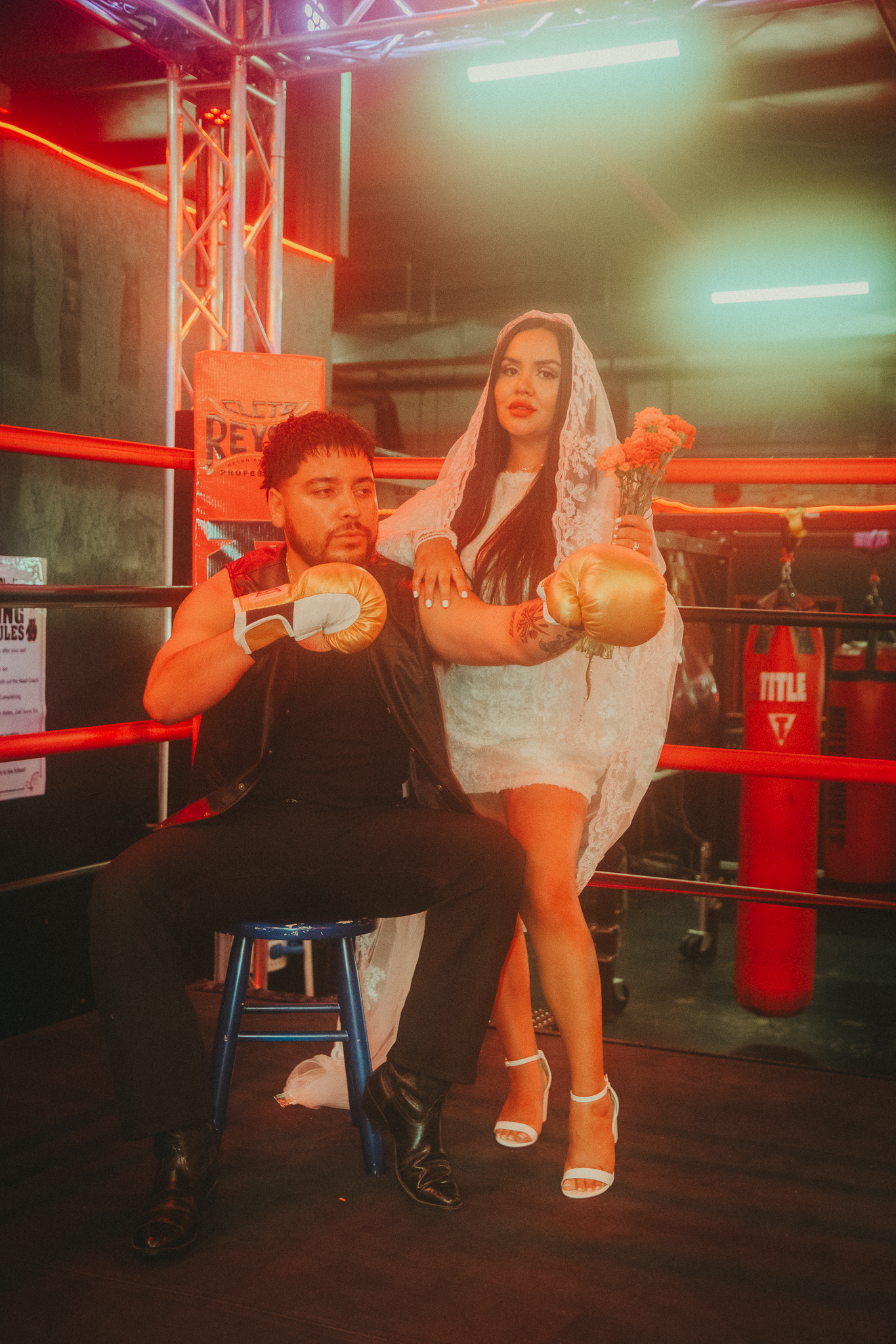 Couple posing in boxing ring, man seated with gold gloves, woman in white dress and veil holding boxing gloves.