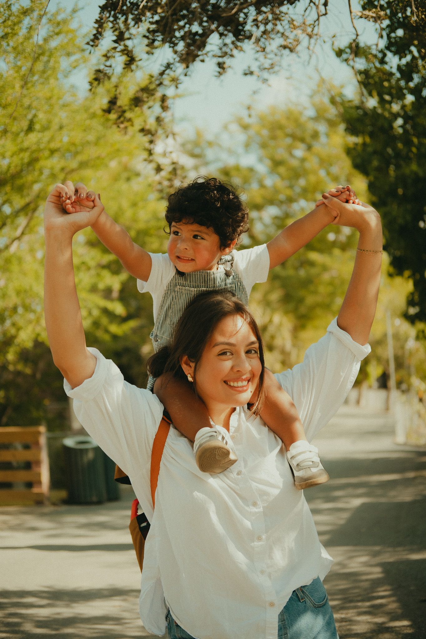 A smiling woman carries a happy child on her shoulders outdoors, both with arms raised in a sunny park.