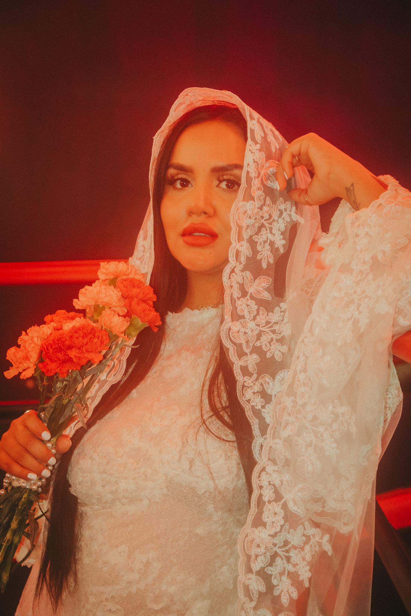 A bride in white lace dress and veil holds orange carnations under dramatic red lighting.