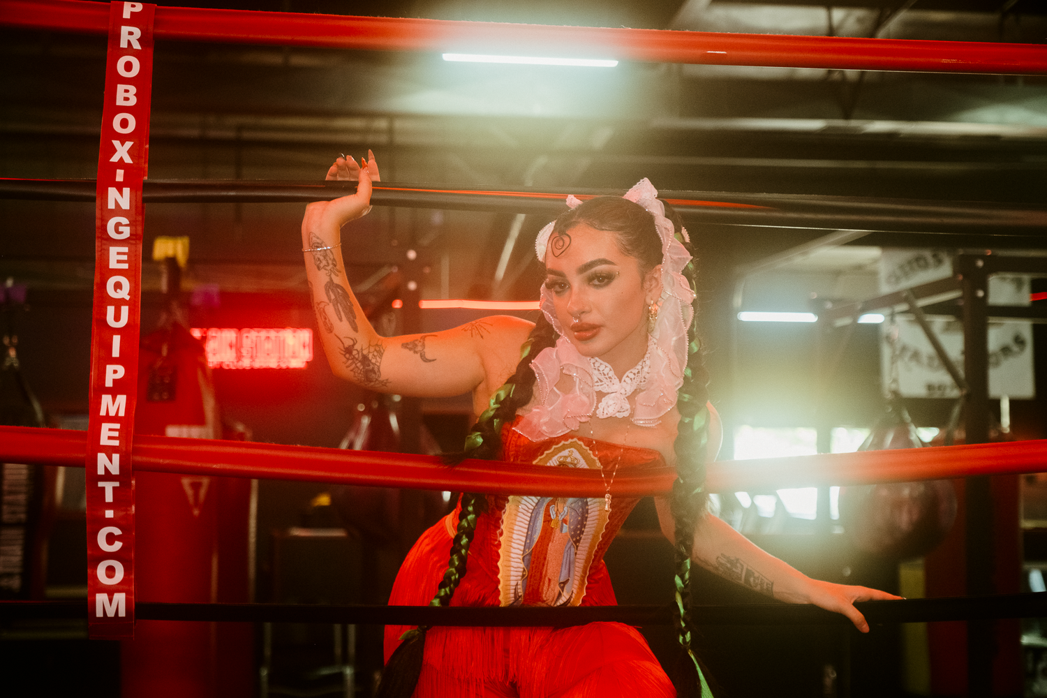 Woman in colorful dress poses confidently in a boxing ring with red ropes and gym equipment in background.