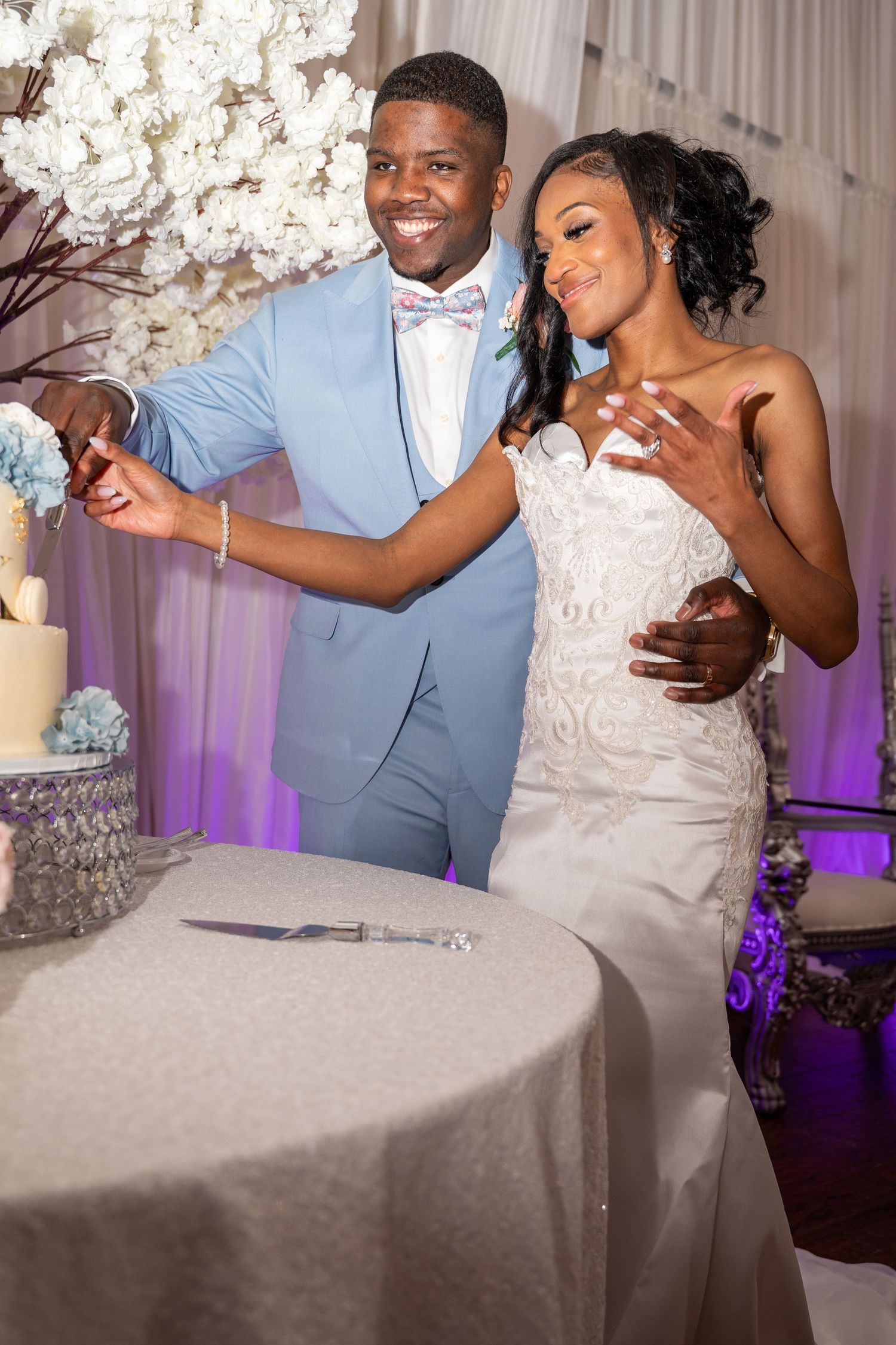 Bride and groom in light blue suit smiling while cutting tiered wedding cake together.