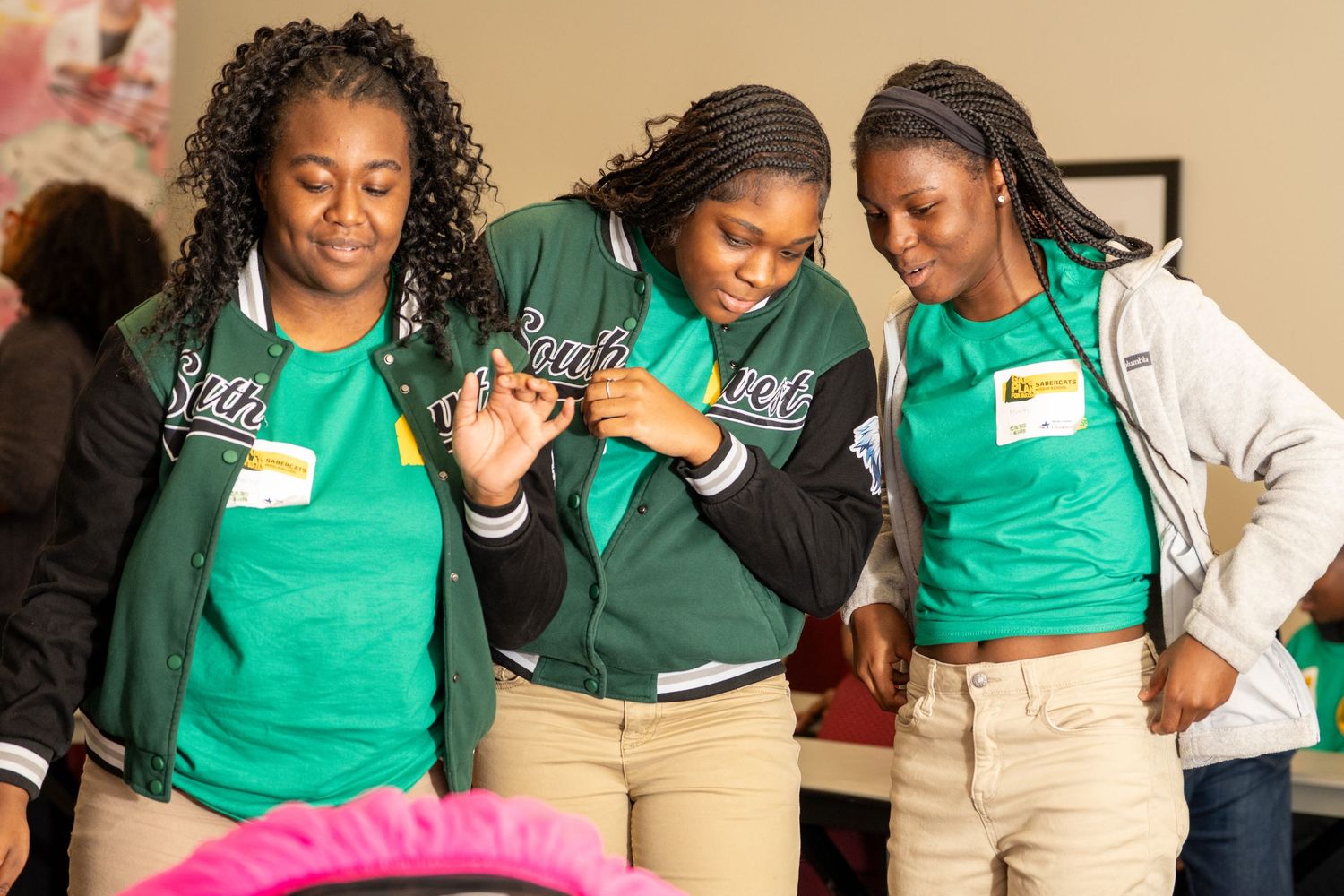 Three teenage girls in green team jackets and t-shirts laugh together at a youth program event.