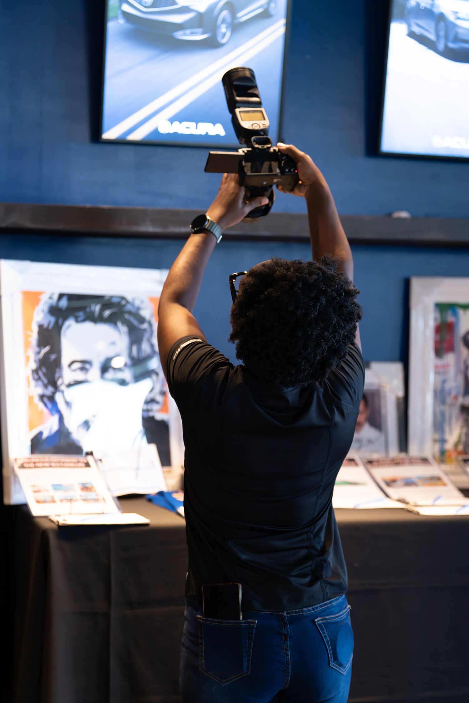 Black photographer holding camera overhead to photograph framed artwork at indoor gallery event. bratcher photography and houston