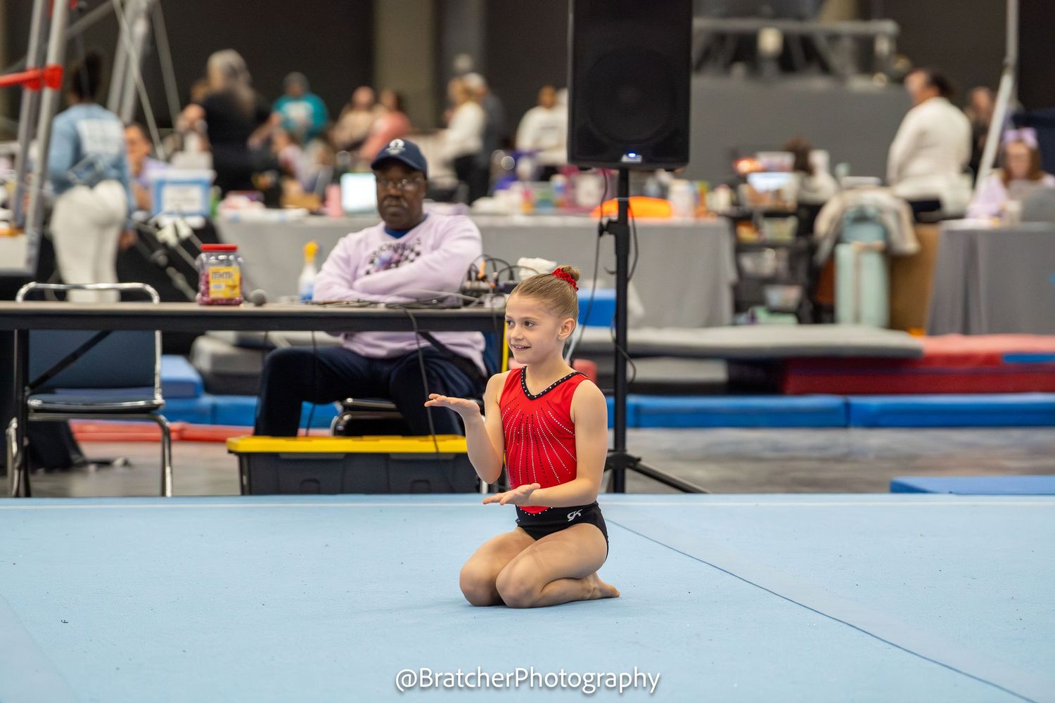 Young female gymnast sits on floor mat smiling after completing routine at indoor gymnastics meet.