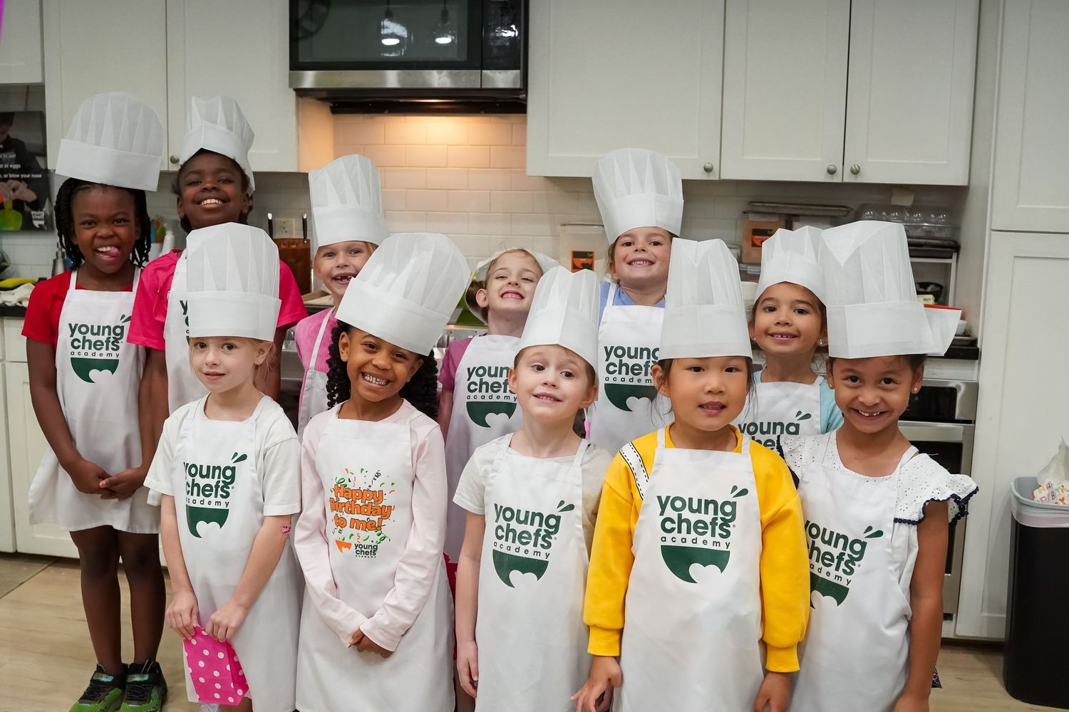Group of young children wearing white chef hats and aprons at a Young Chefs cooking class.