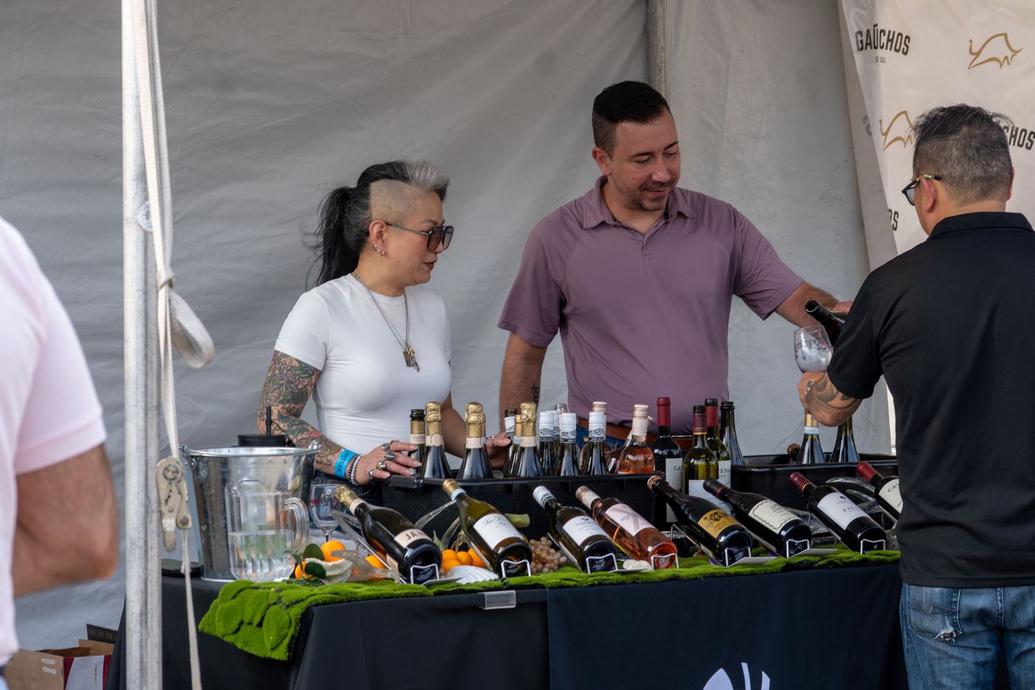 Two vendors display wine bottles at an outdoor festival tasting booth under a white tent.