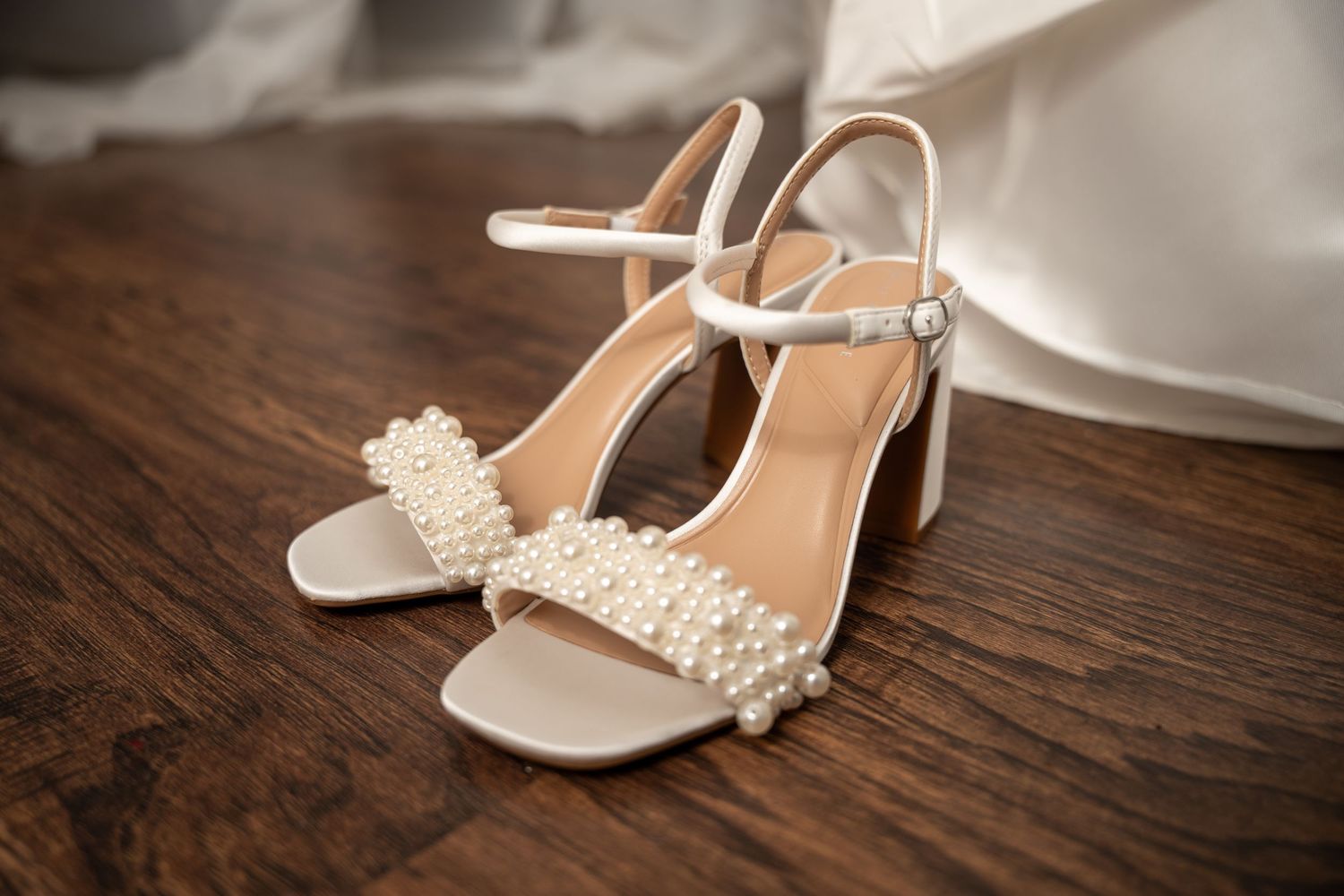 White pearl-embellished block heel bridal sandals on wooden floor before wedding.