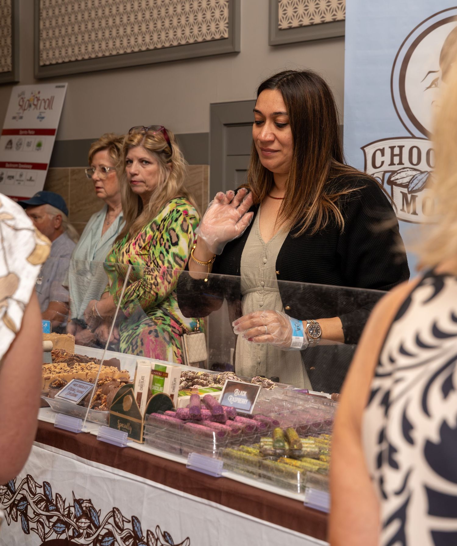 Woman gestures while customers browse chocolate display case at Choco M artisan chocolate shop.