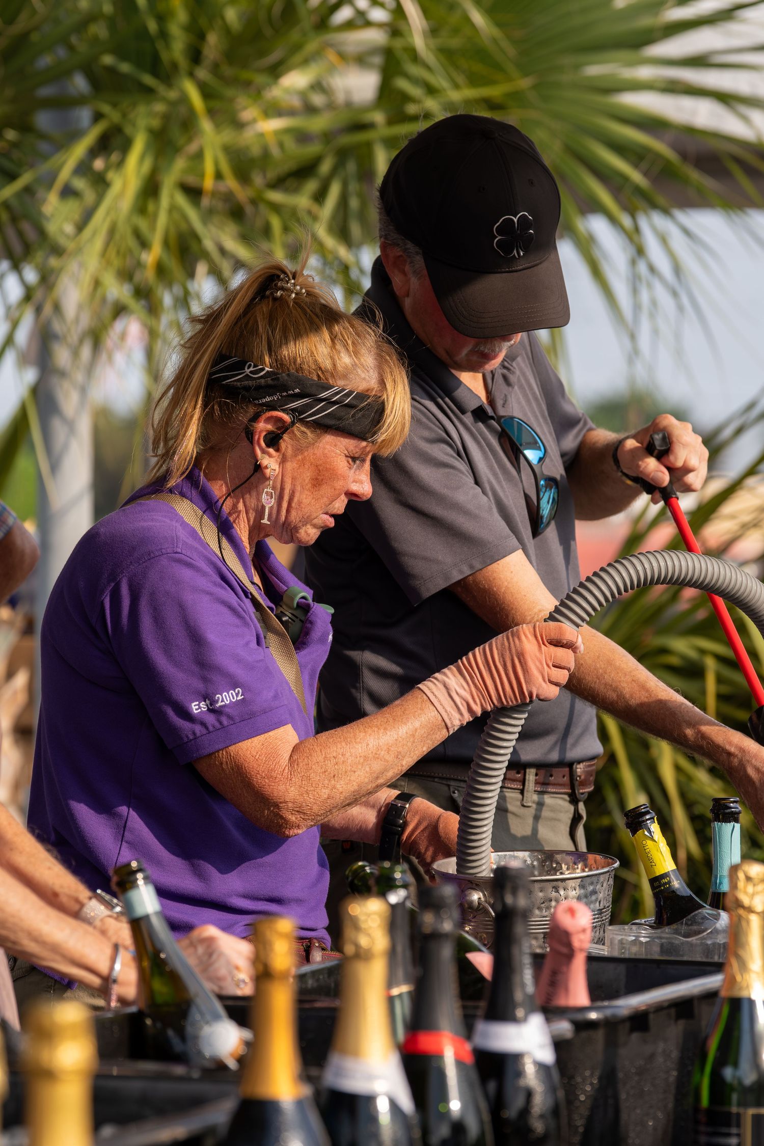 Two workers managing champagne and wine bottles at an outdoor festival beverage station.