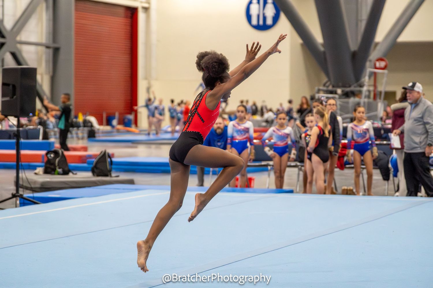 Young gymnast leaps gracefully during floor exercise at indoor gymnastics competition wearing red leotard.
