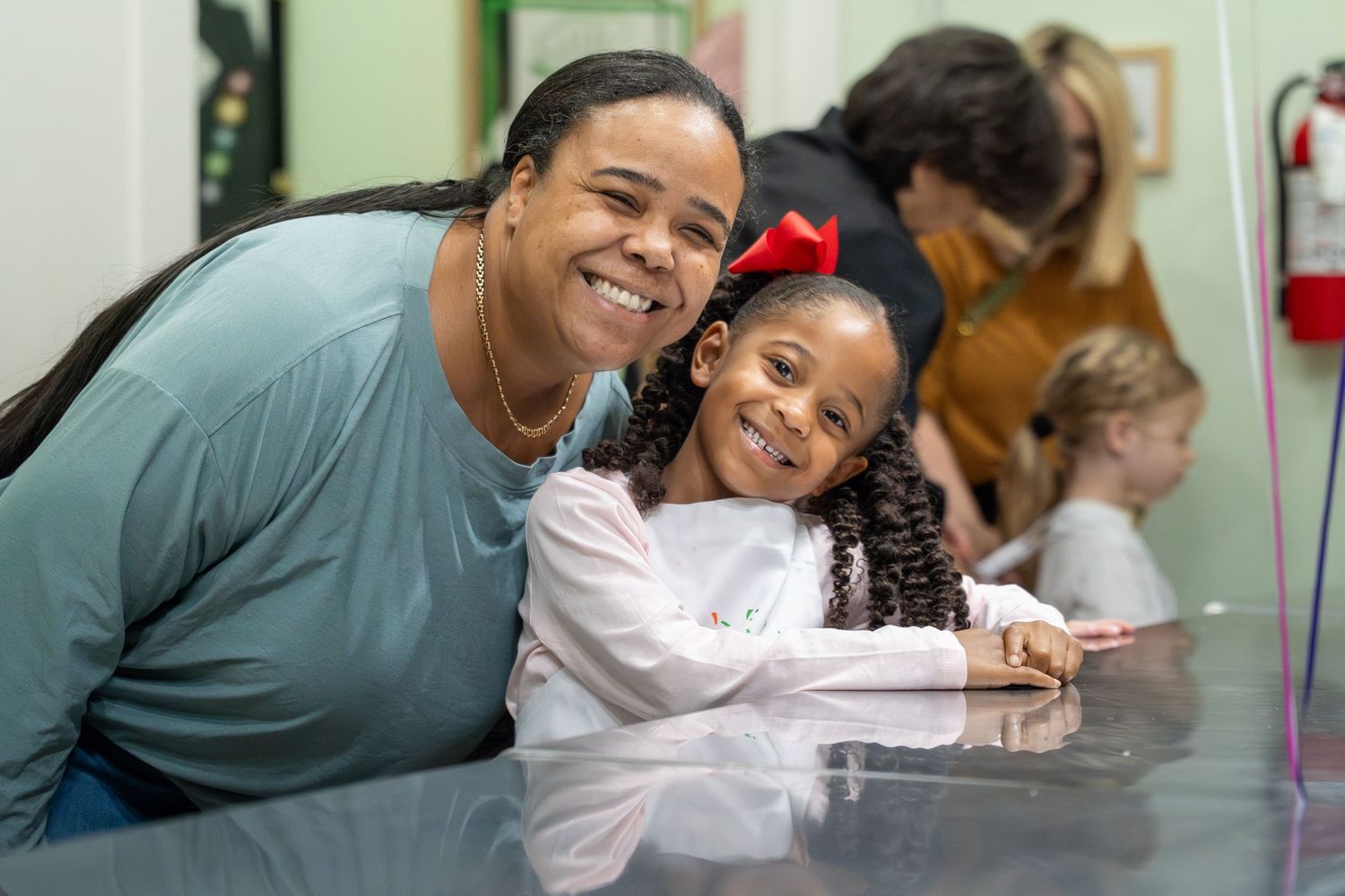 Smiling woman and young girl with red bow posing together at a table indoors.