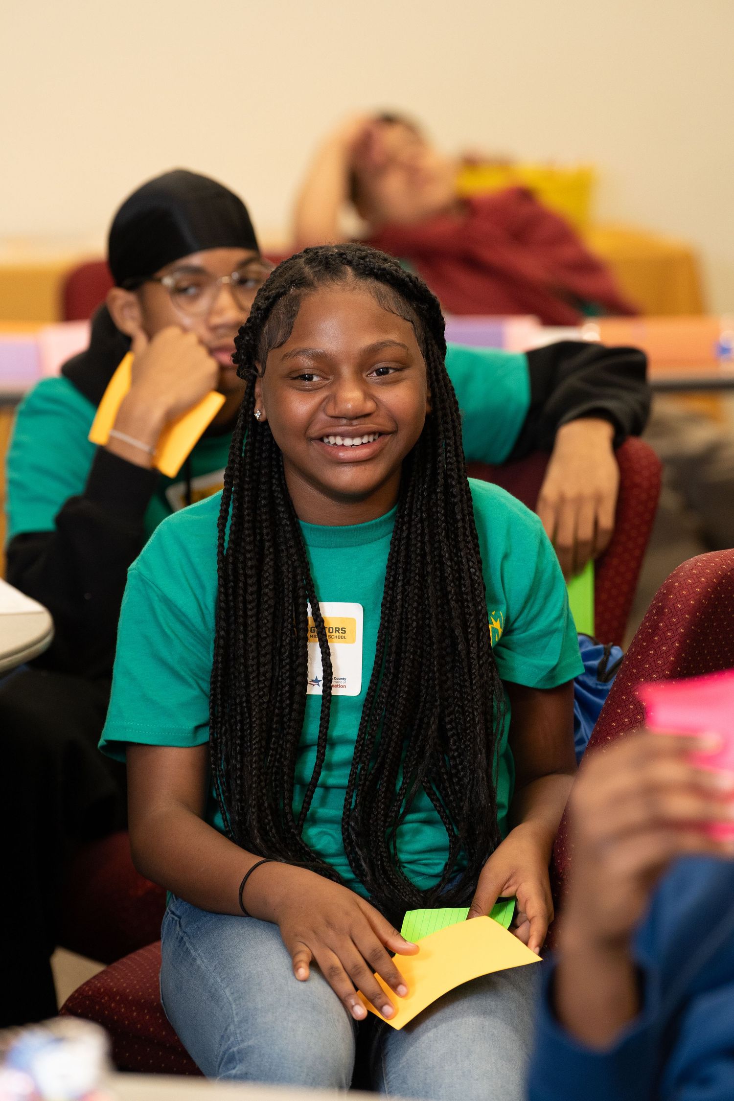 Smiling teenage girl in green t-shirt holds colorful papers at a youth educational workshop.