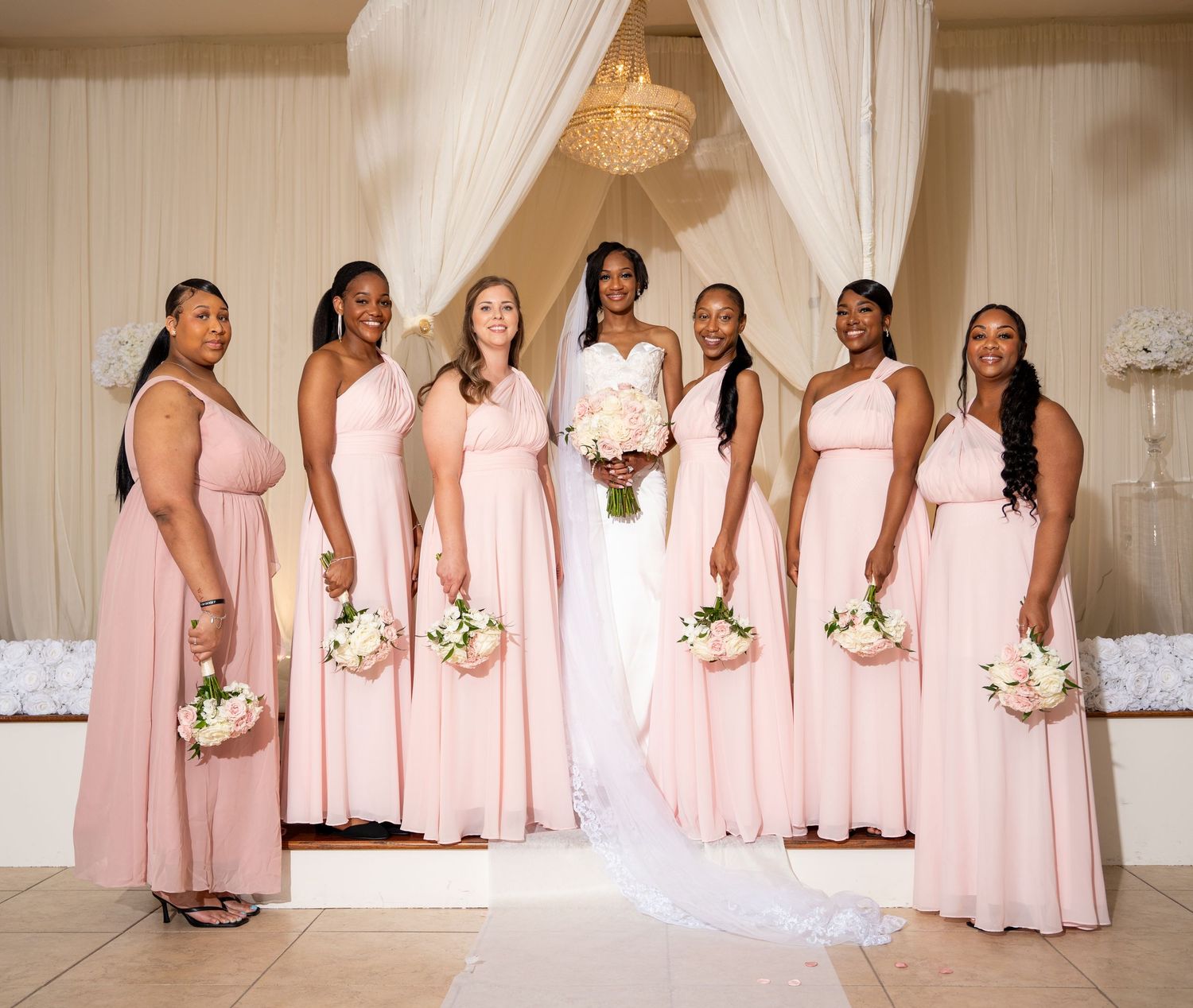 Bride surrounded by six bridesmaids in blush pink dresses holding white bouquets at wedding venue.