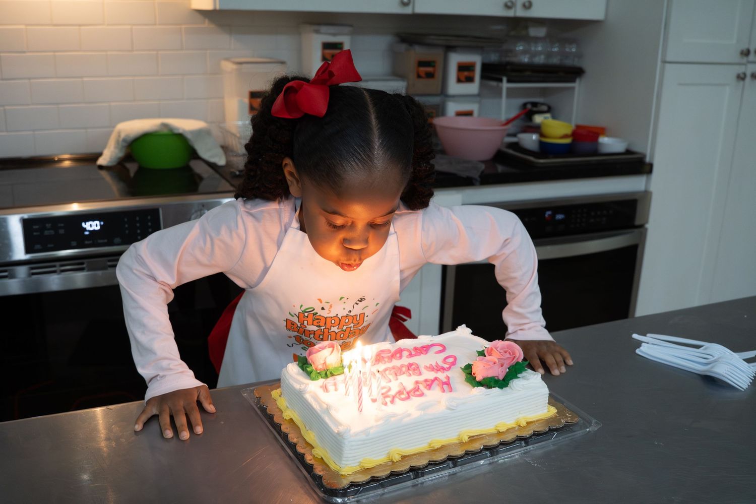 Young girl blows out candles on a decorated birthday cake in a modern white kitchen.