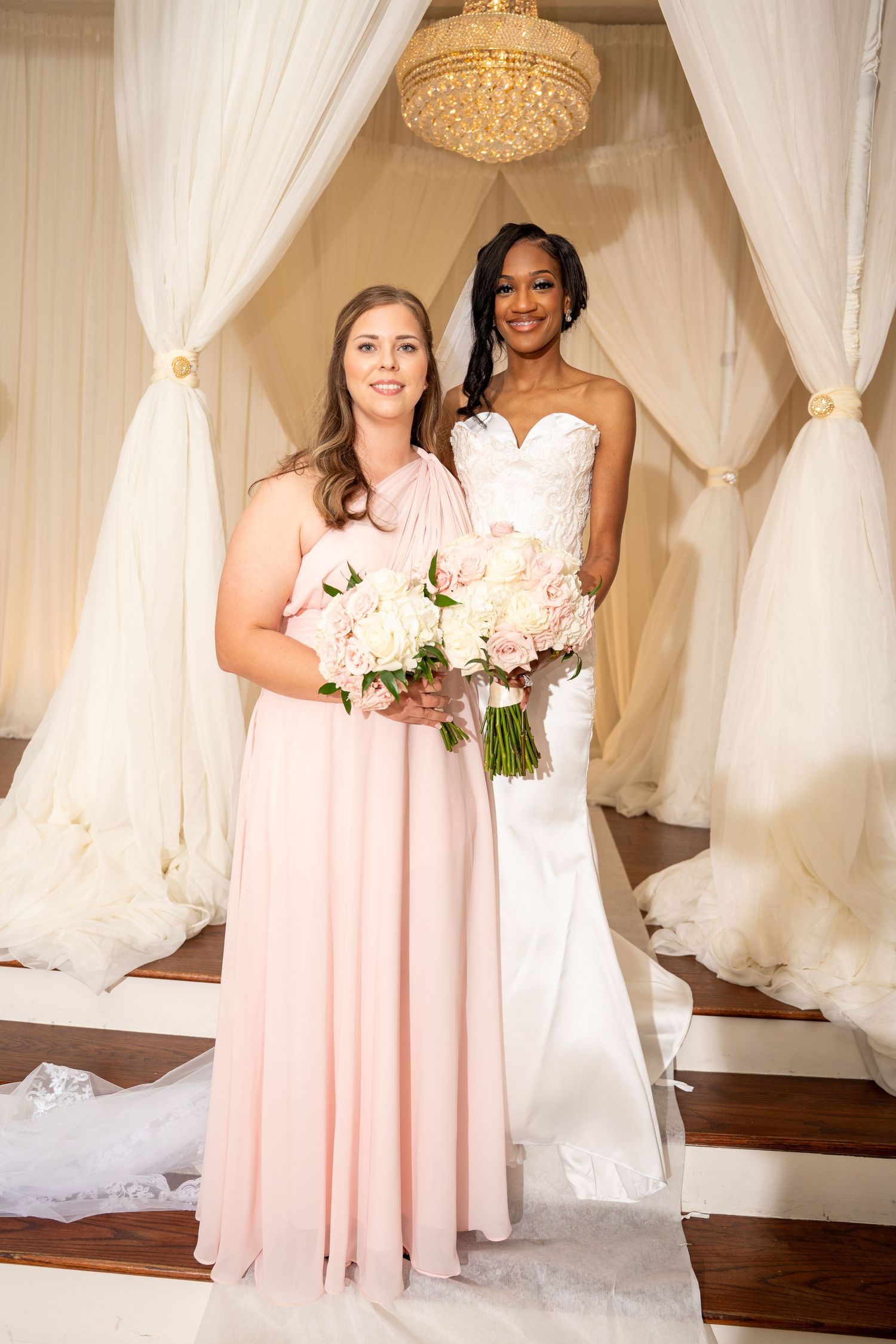 Bride in white lace gown poses with bridesmaid in blush pink dress at wedding ceremony.