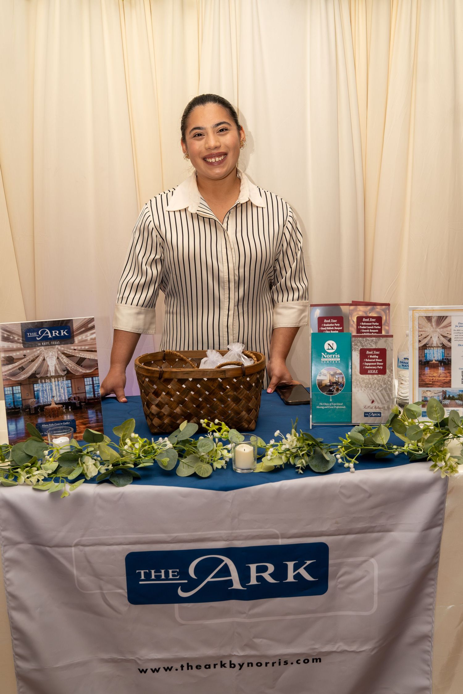 Smiling woman stands at The Ark by Norris event venue booth with greenery and brochure display.