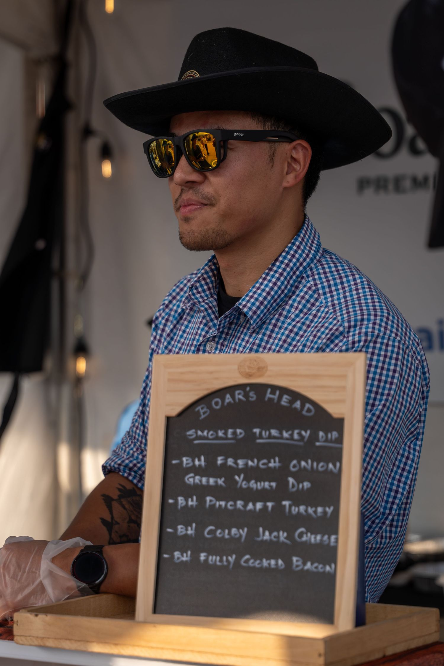 Man in black cowboy hat and sunglasses stands beside a Boars Head chalkboard menu at food festival.
