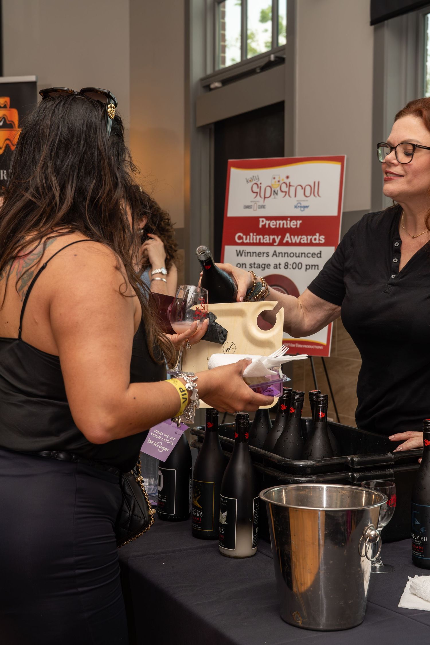 Woman receives wine pour at Sip and Stroll Premier Culinary Awards event from smiling server at bottle-lined table.