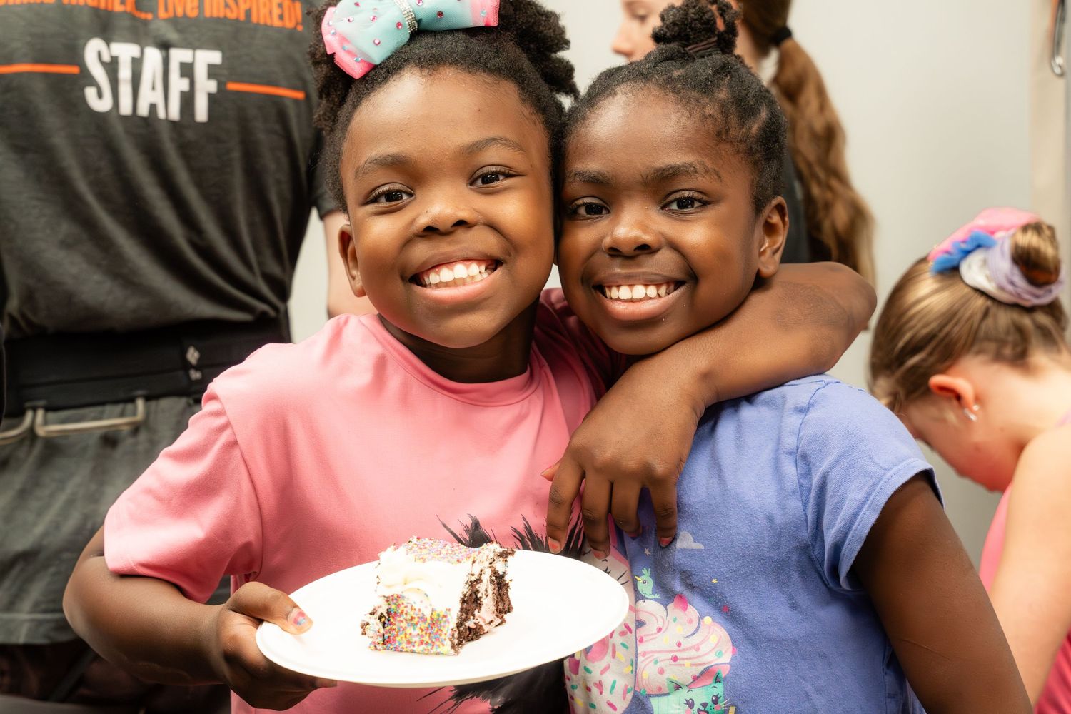 Two smiling girls hugging and holding a plate of birthday cake at a party.