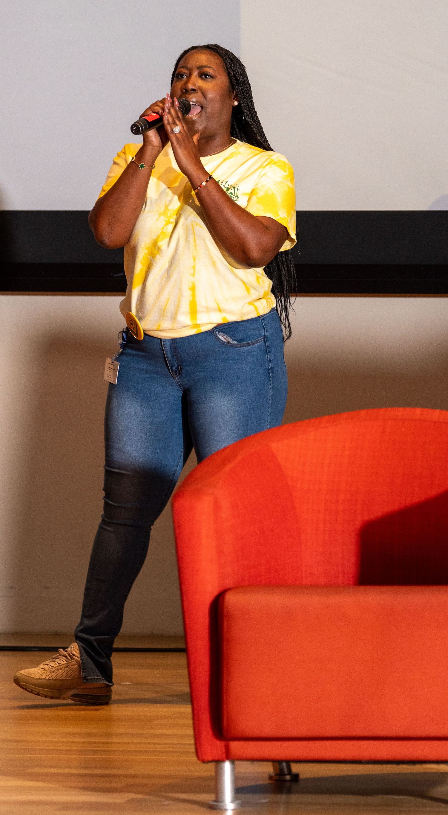 Young woman in yellow tie-dye shirt sings into microphone on stage beside a red chair.