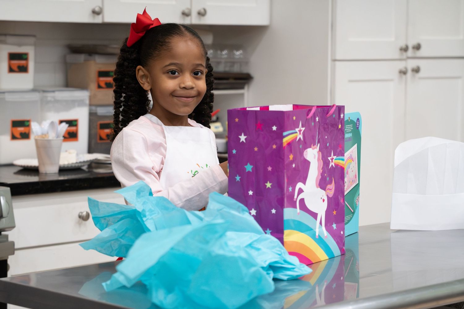 Young girl smiles beside a unicorn gift bag with teal tissue paper at a birthday celebration.