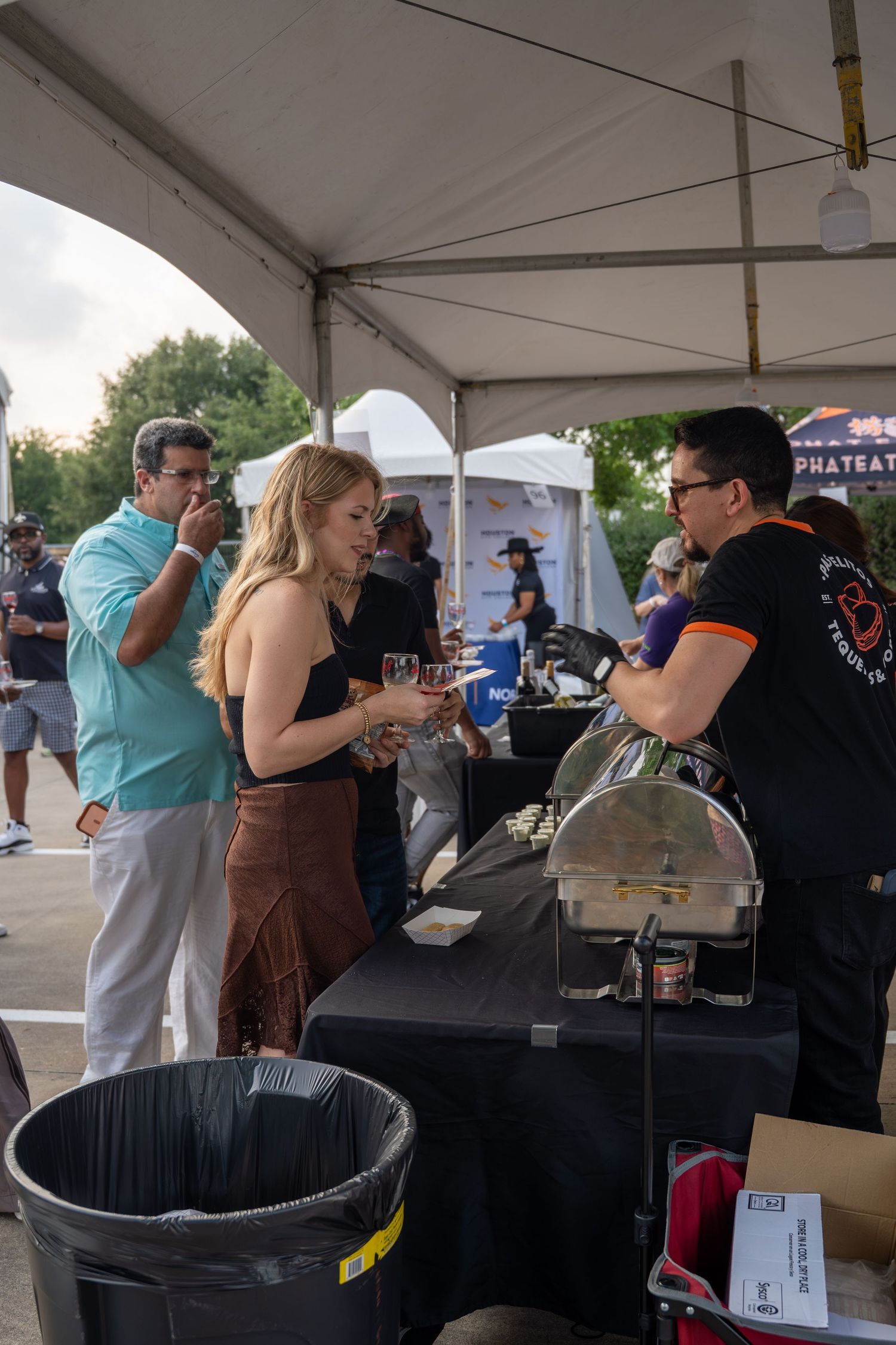 Couple samples food from chafing dish at outdoor culinary festival booth under white tent.