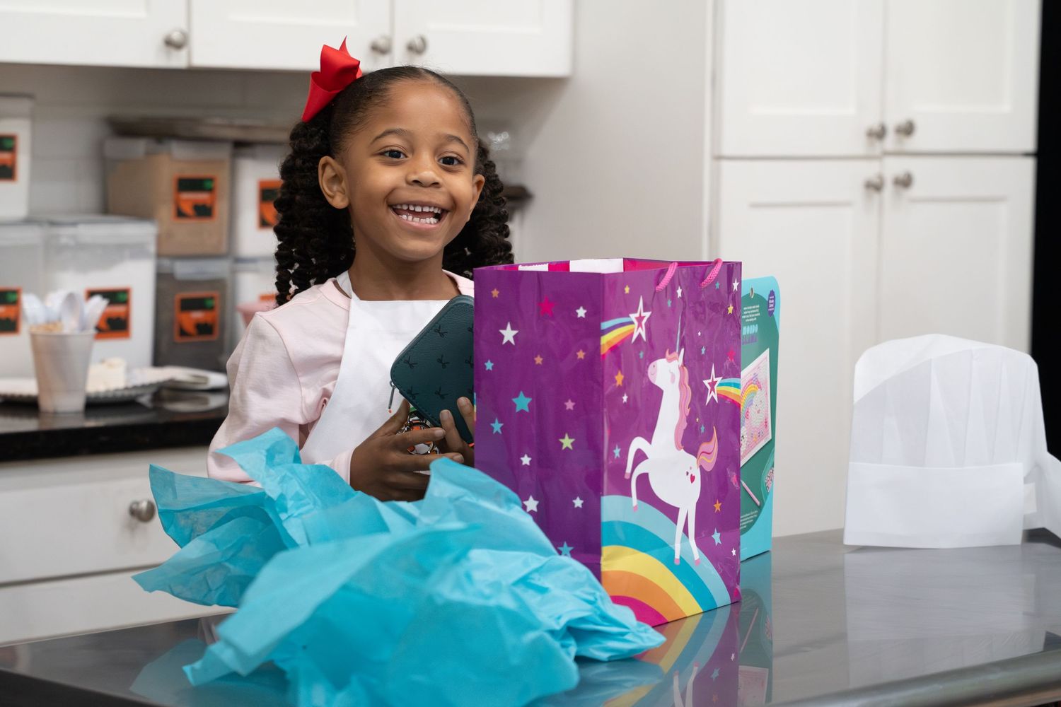 Smiling young Black girl celebrating birthday with colorful unicorn gift bags and teal tissue paper on kitchen counter. bratcher photography