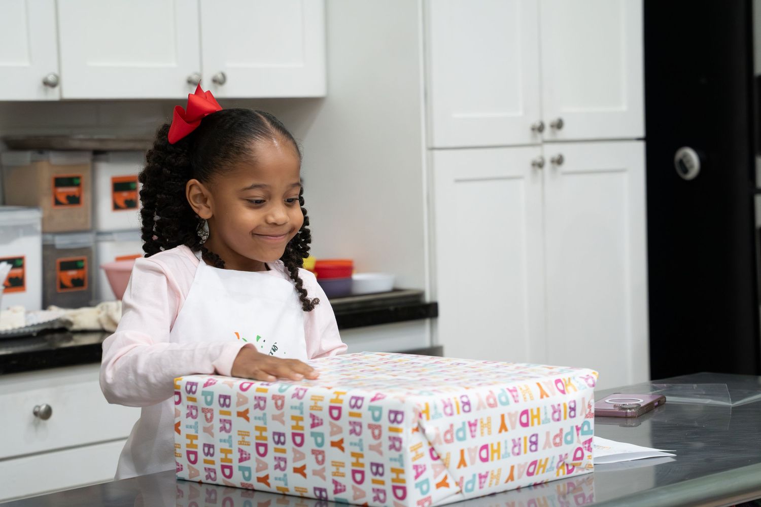 Young girl smiling while opening a colorful alphabet-wrapped birthday gift on a kitchen counter.