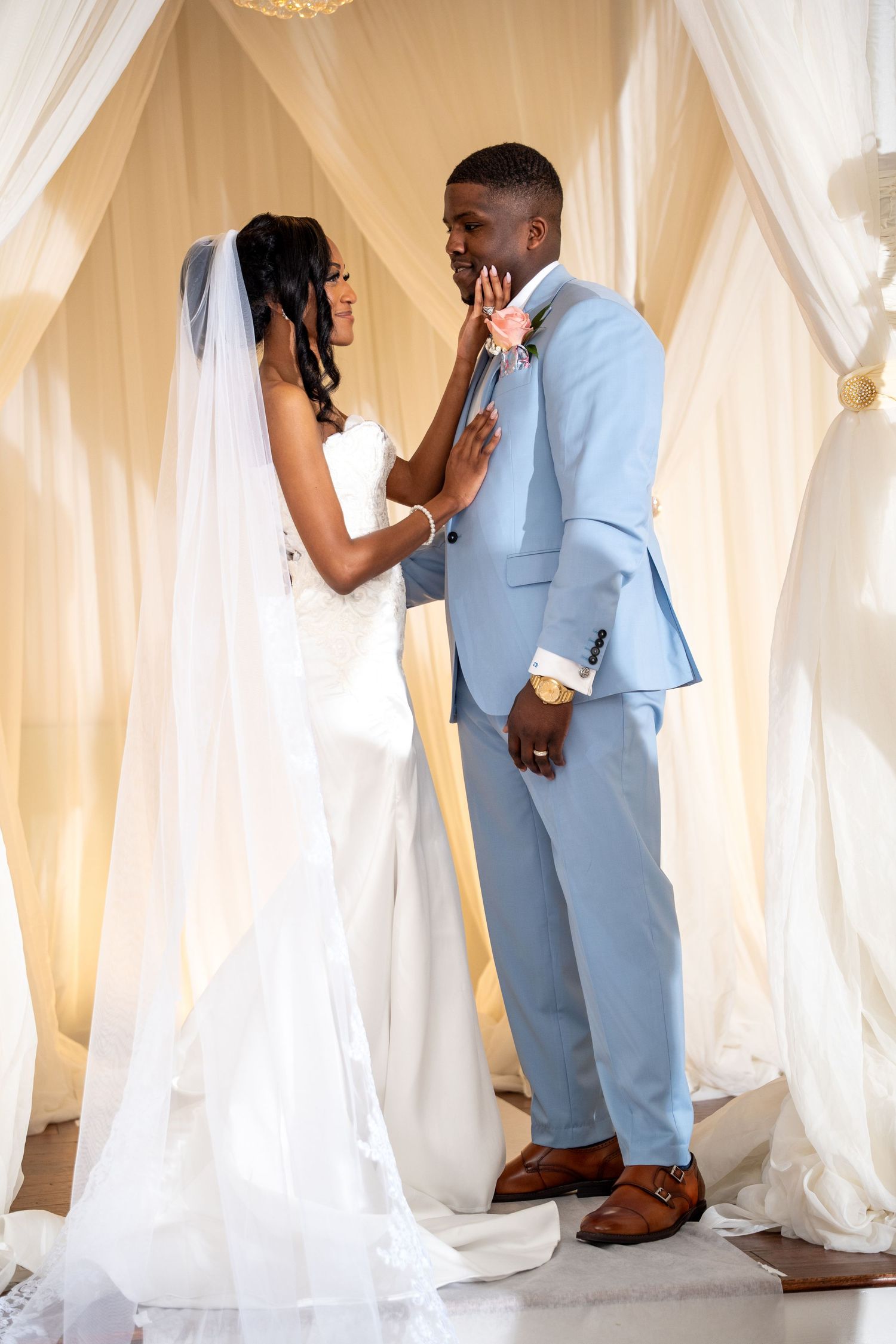 Bride and groom in light blue suit sharing tender moment under elegant draped wedding arch.