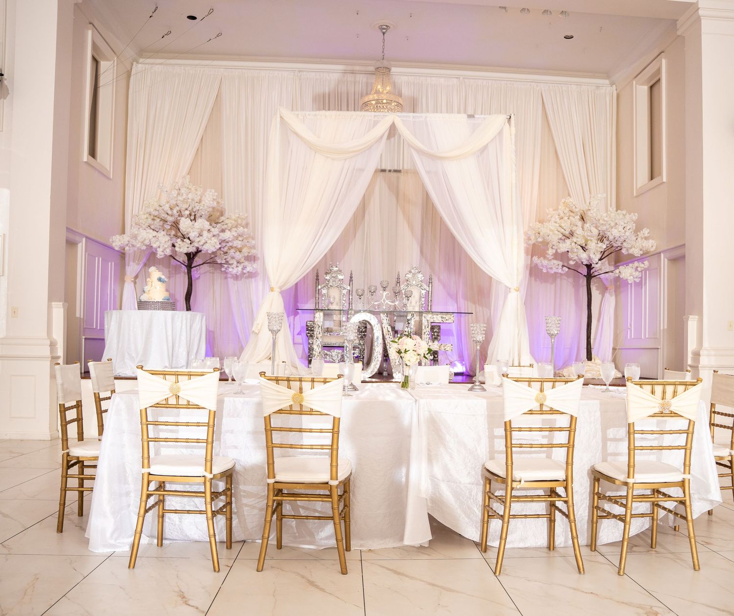 Elegant wedding reception hall with white draping, gold chiavari chairs, and cherry blossom centerpieces.