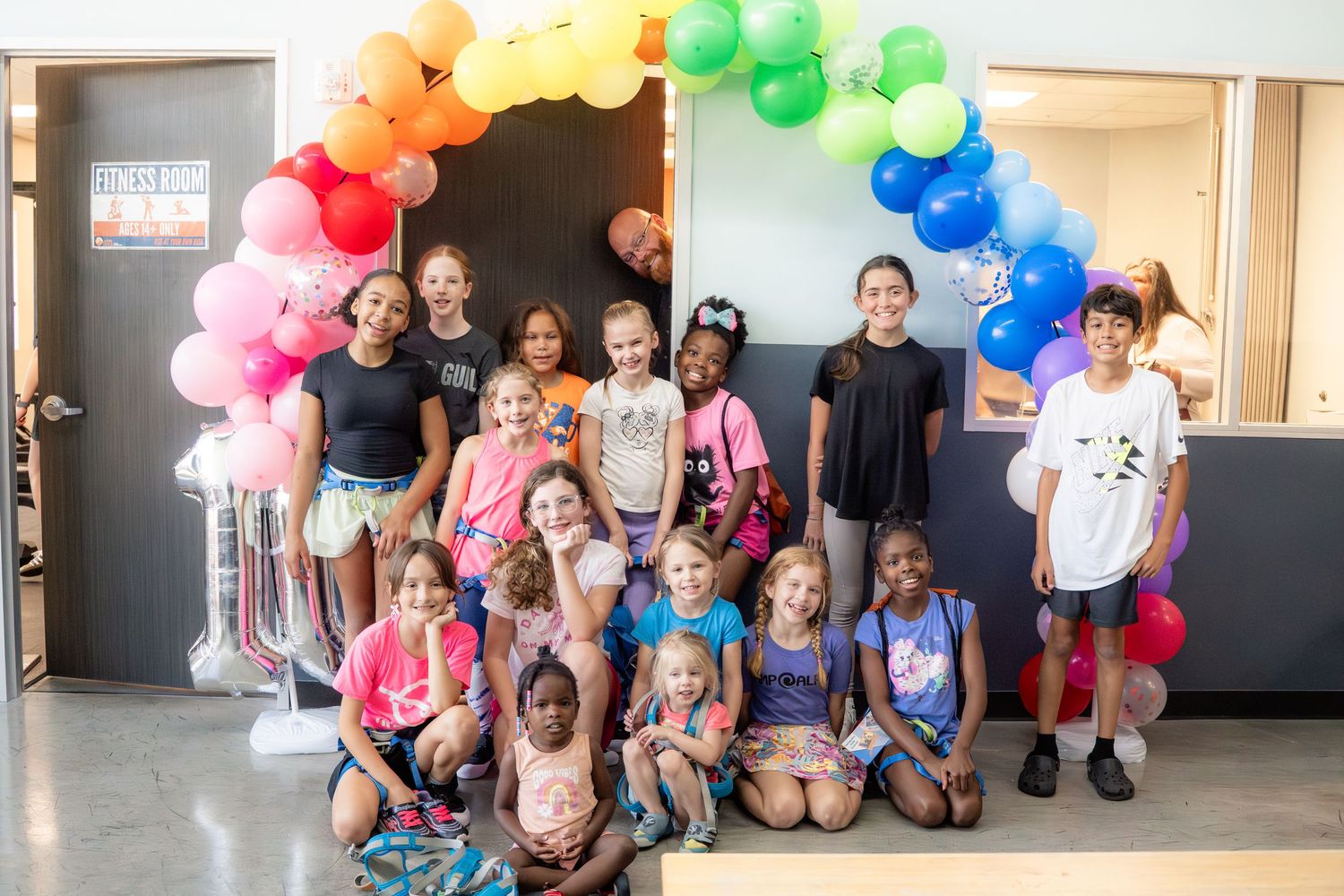 Group of diverse children posing under colorful rainbow balloon arch indoors.