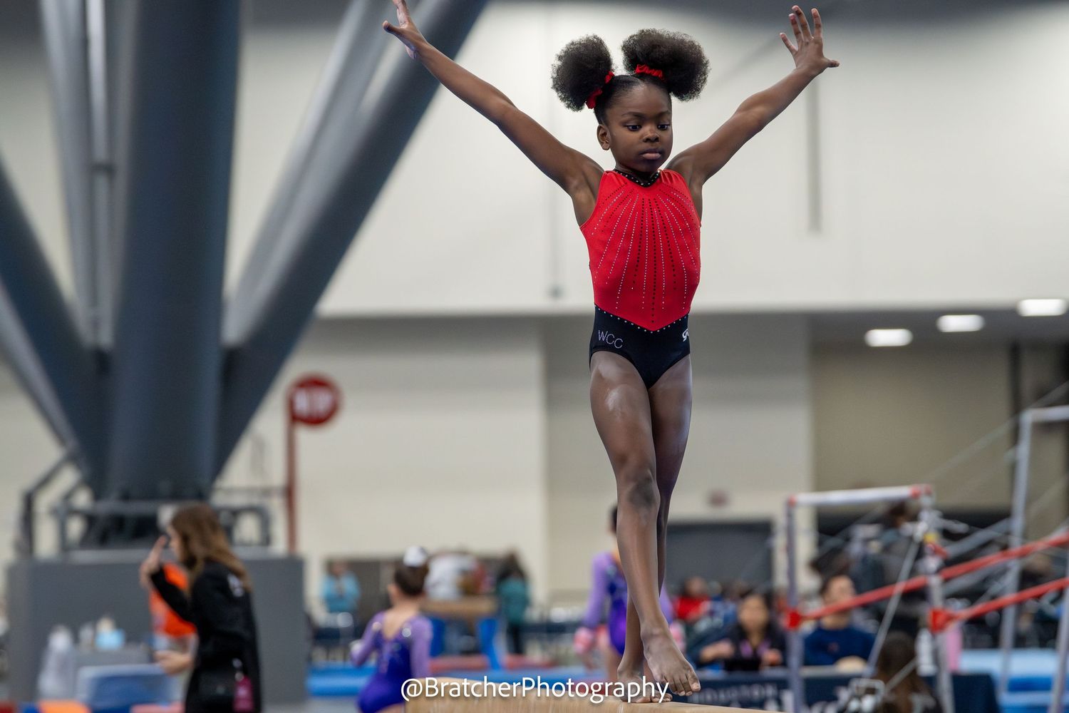 Young Black gymnast balances on beam with arms raised at indoor gymnastics competition in red leotard.