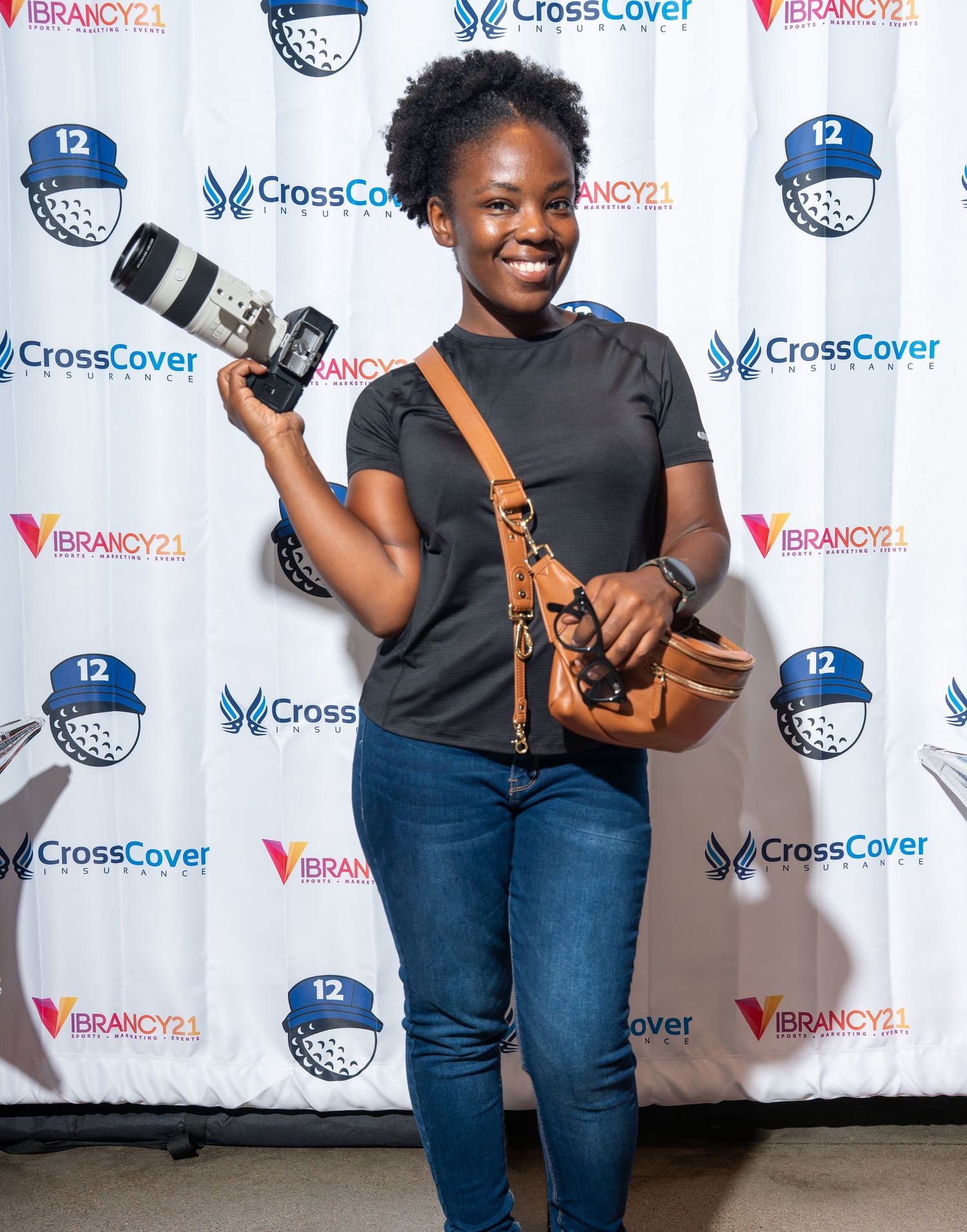 Young woman smiling while holding a camera at a CrossCover Vibrancy21 event backdrop.