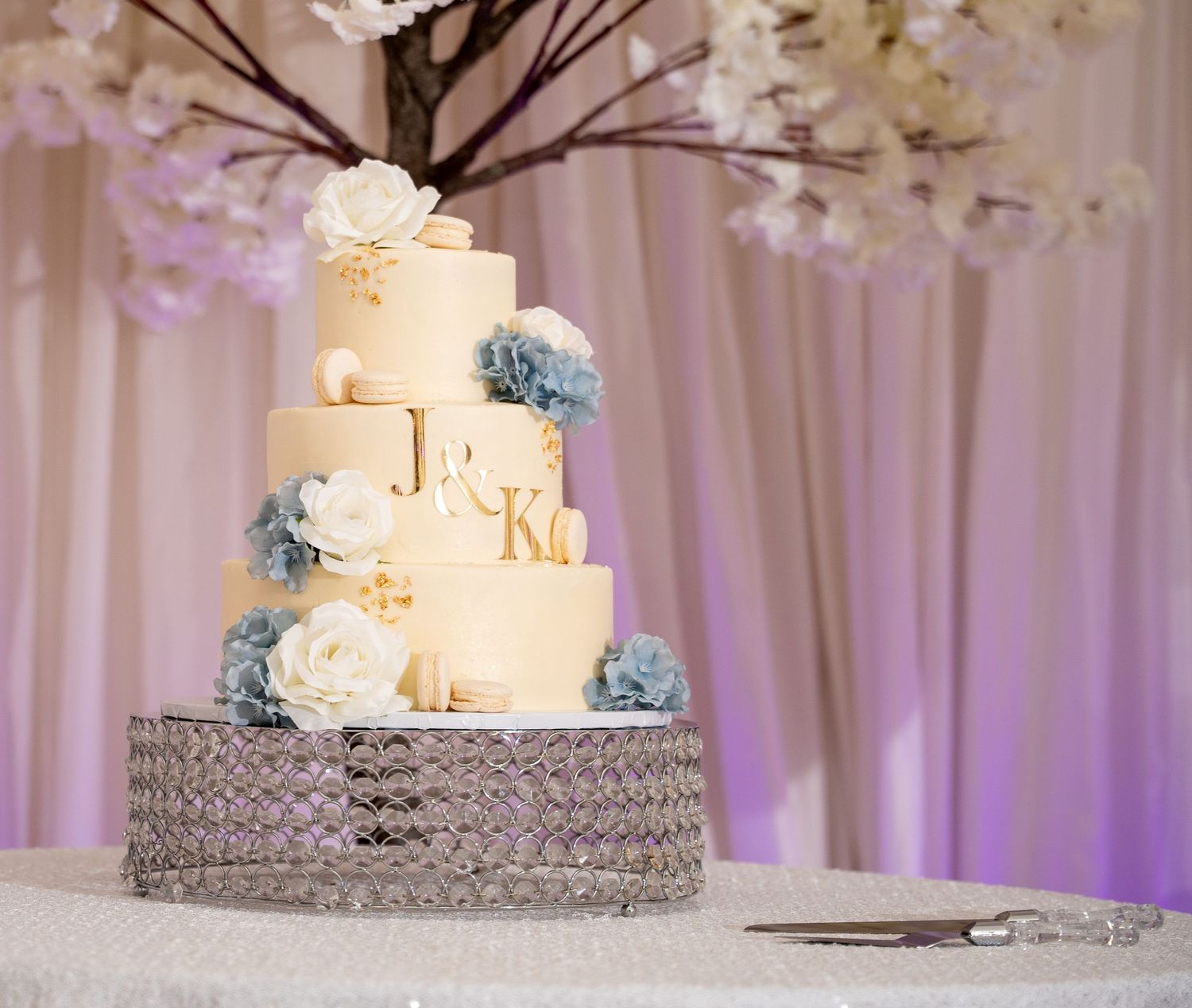 Four-tier ivory wedding cake decorated with blue and white flowers on a crystal cake stand.