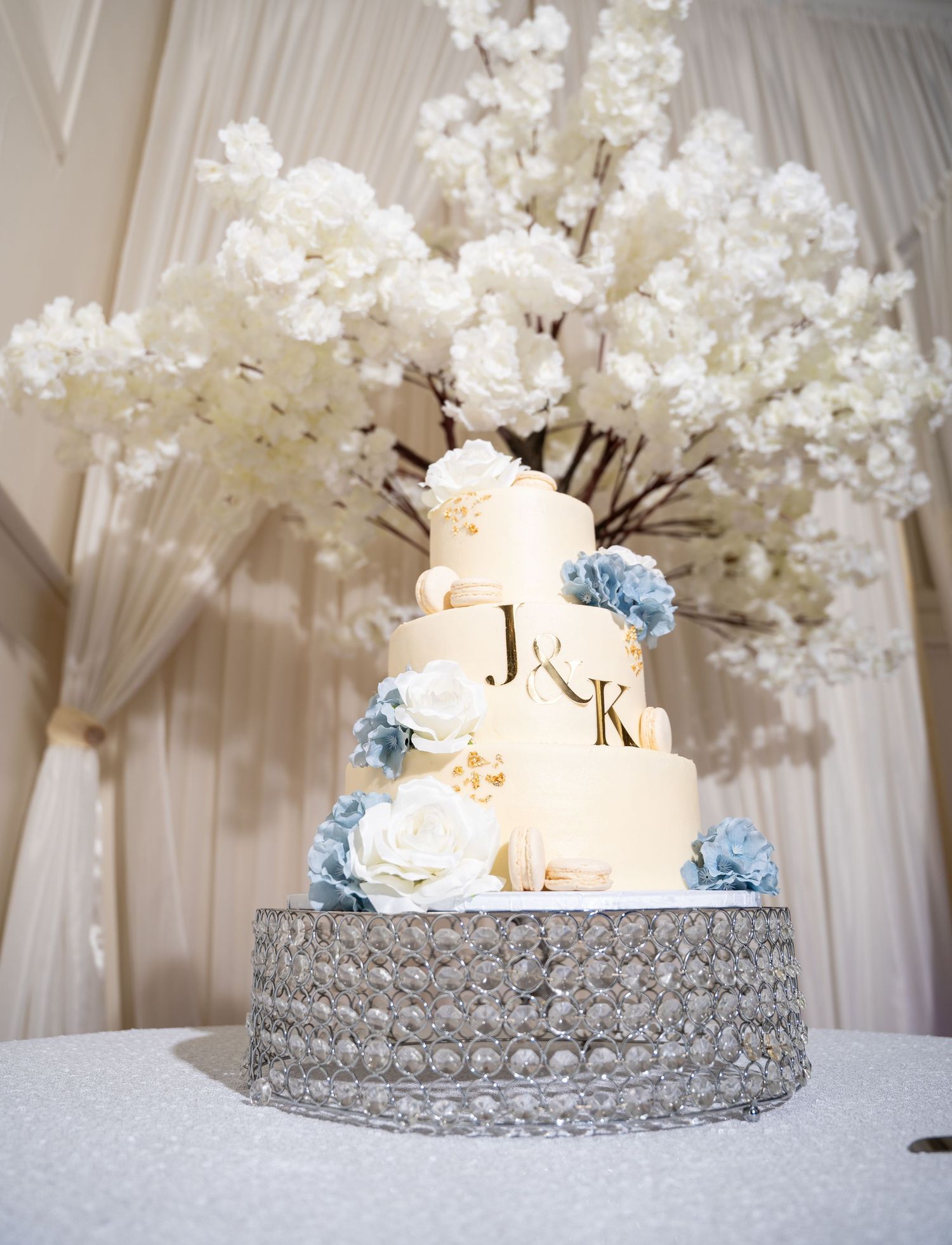 Four-tier white wedding cake with blue floral accents on crystal stand with cherry blossom backdrop.