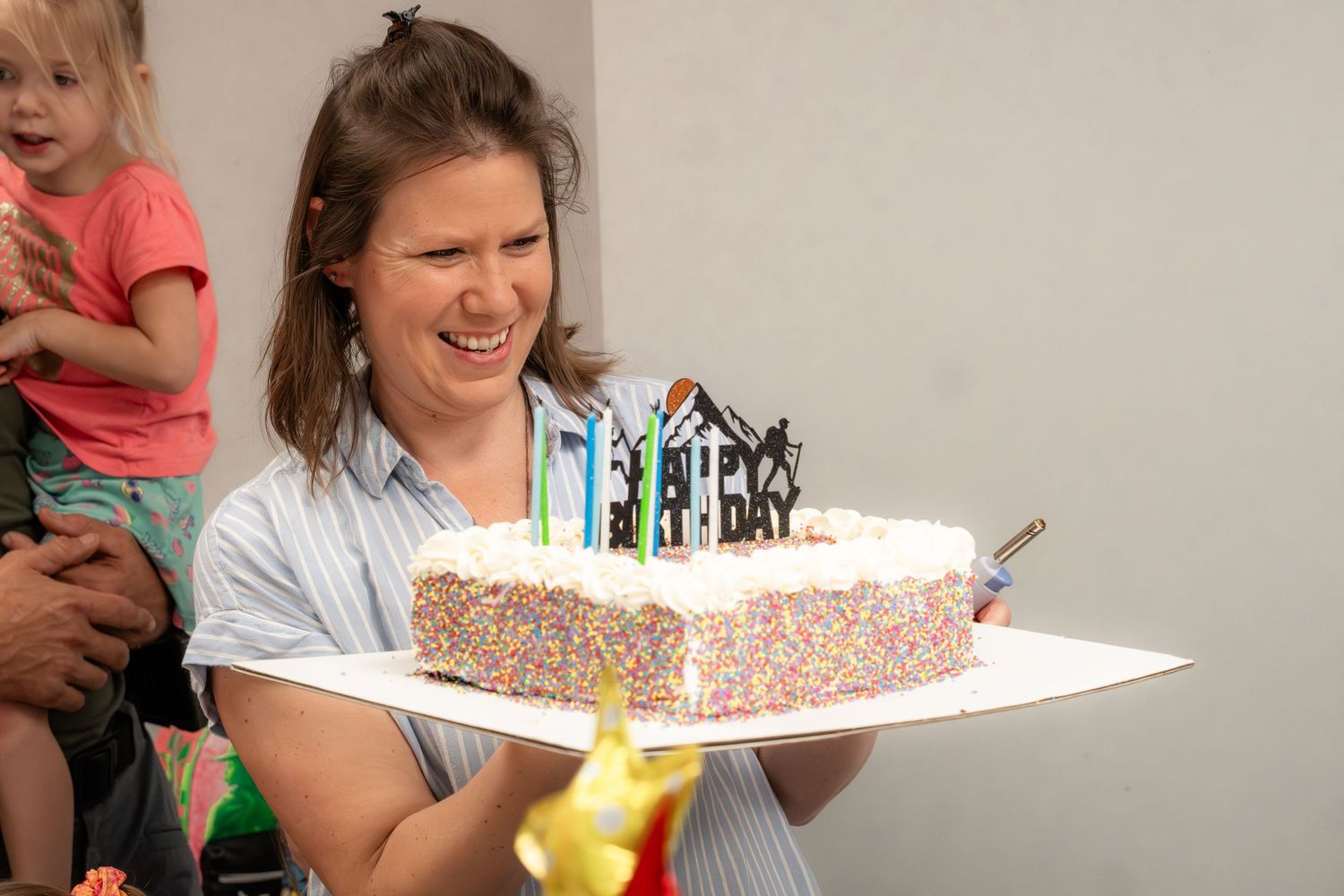 Woman smiling while holding a colorful sprinkle birthday cake with candles indoors.