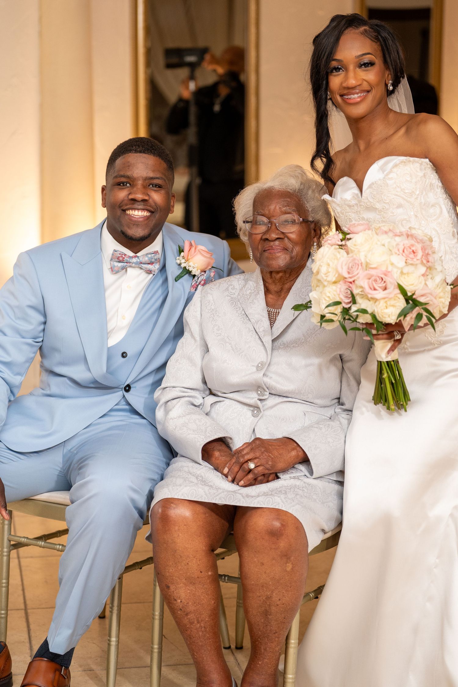 Groom in light blue suit with bride in white lace gown posing with elderly woman at wedding reception.