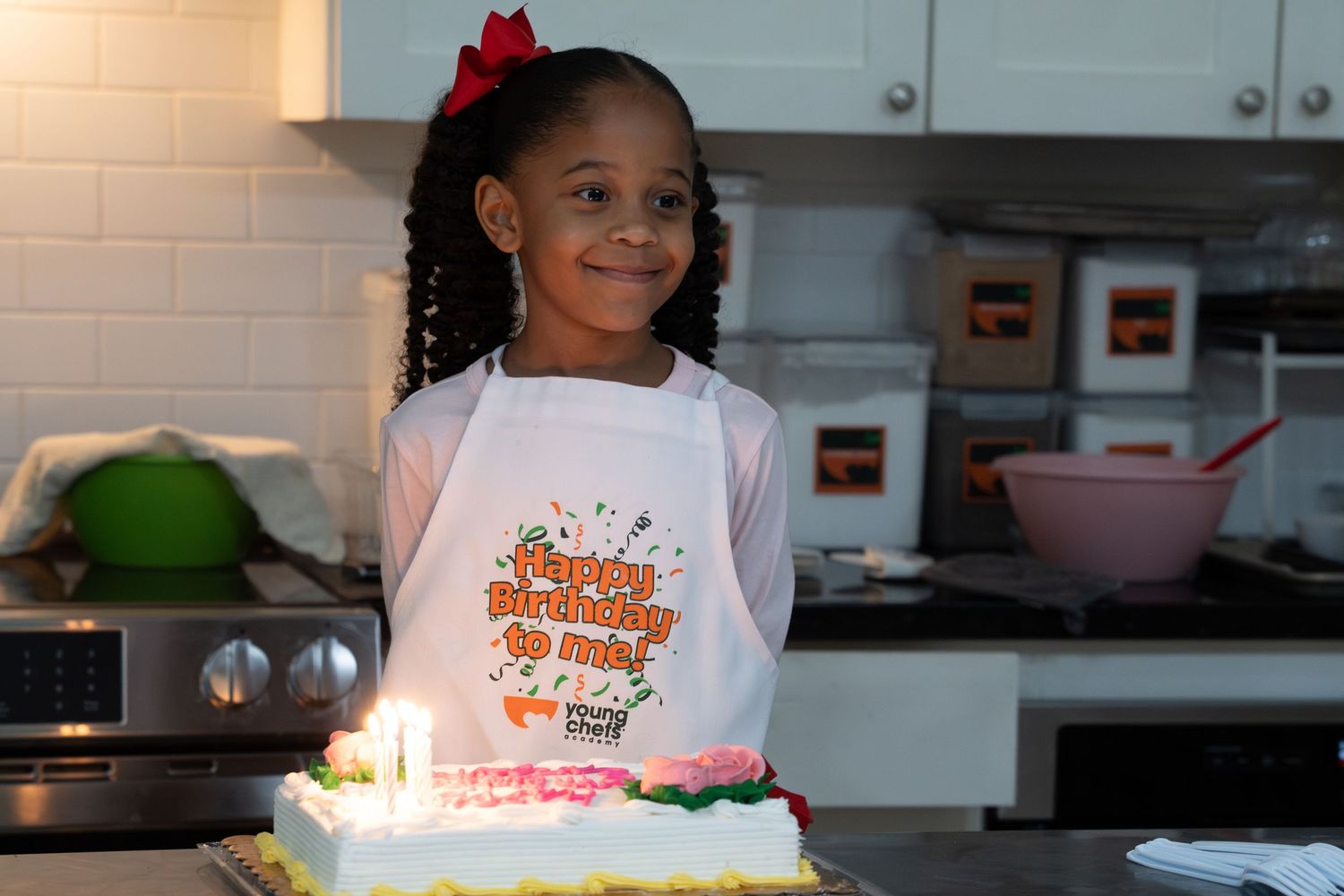 Smiling girl poses in a birthday apron beside a decorated cake with lit candles in kitchen.