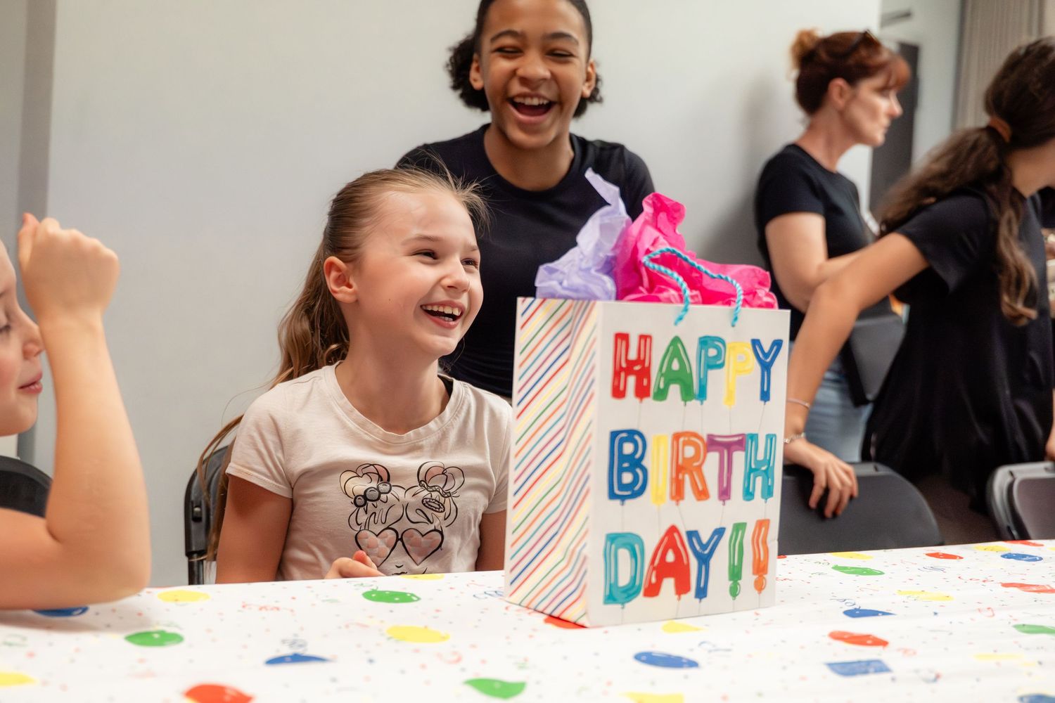 Happy girl laughs beside a Happy Birthday gift bag at a colorful birthday party celebration.