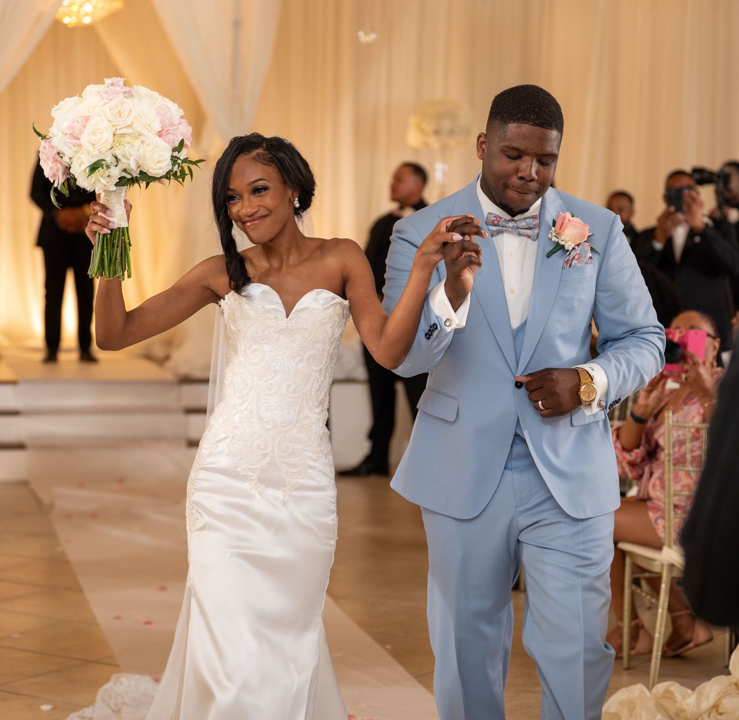 Bride holding bouquet and groom in light blue suit joyfully walking down wedding aisle.