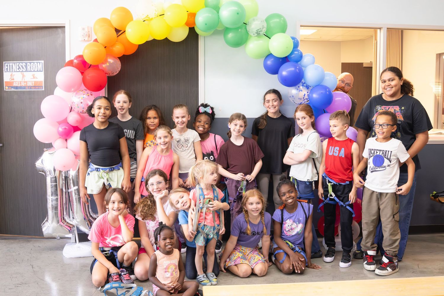 Large group of children posing under a colorful rainbow balloon arch at a party.