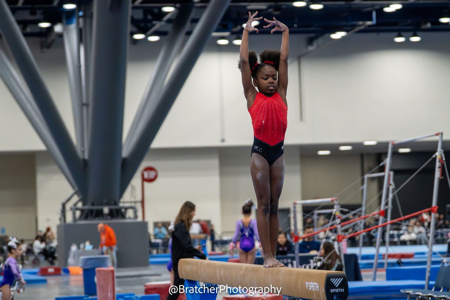 Young gymnast performs balance beam routine with arms raised overhead at indoor gymnastics competition. bratcher photography and houston